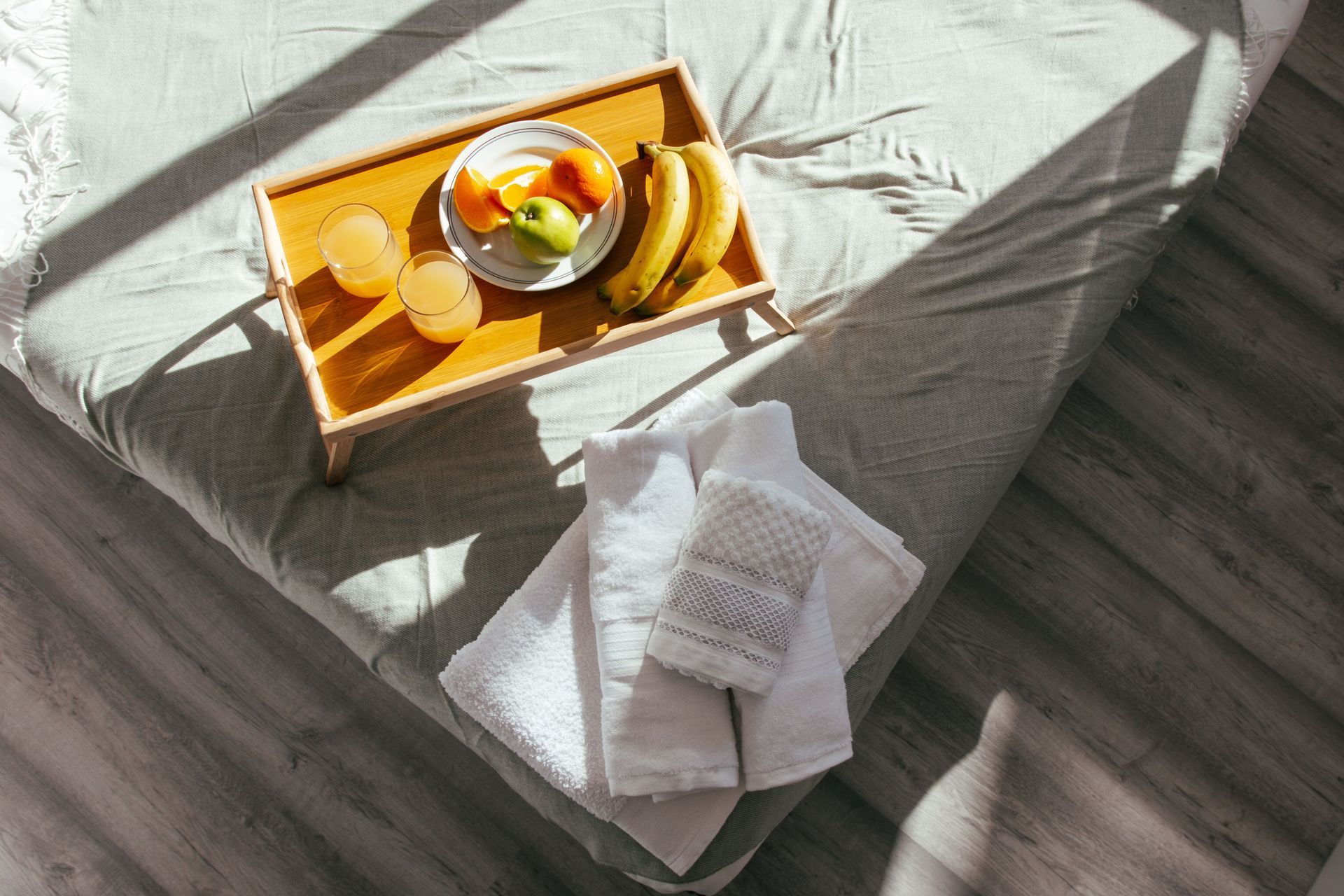 Un vassoio per la colazione sul letto con frutta, una bevanda e asciugamani bianchi piegati, sotto la luce del sole.
