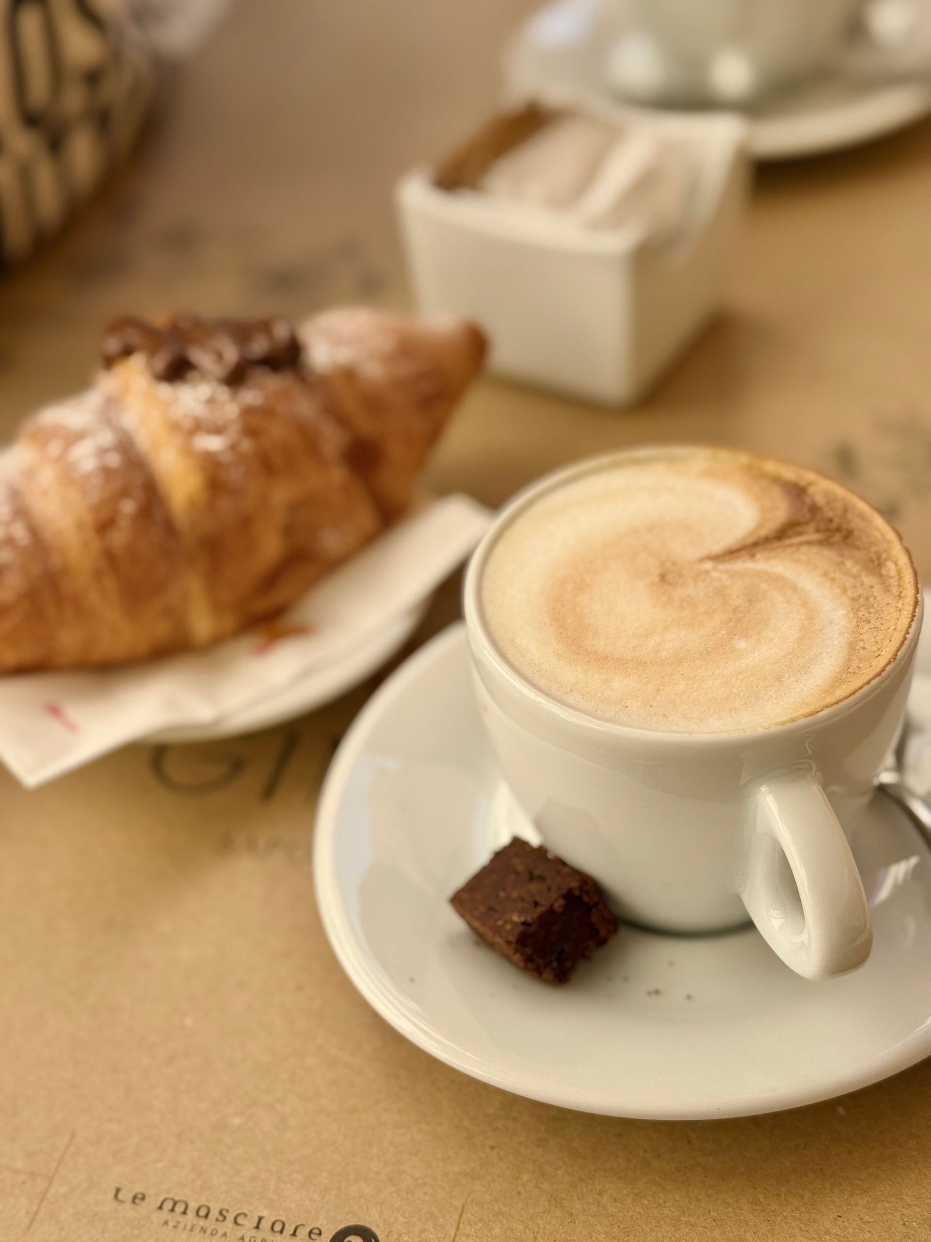 Una tazza di caffè bianca con decorazione di latte art, un croissant e un cioccolatino su un piattino, appoggiati al tavolino di un bar.