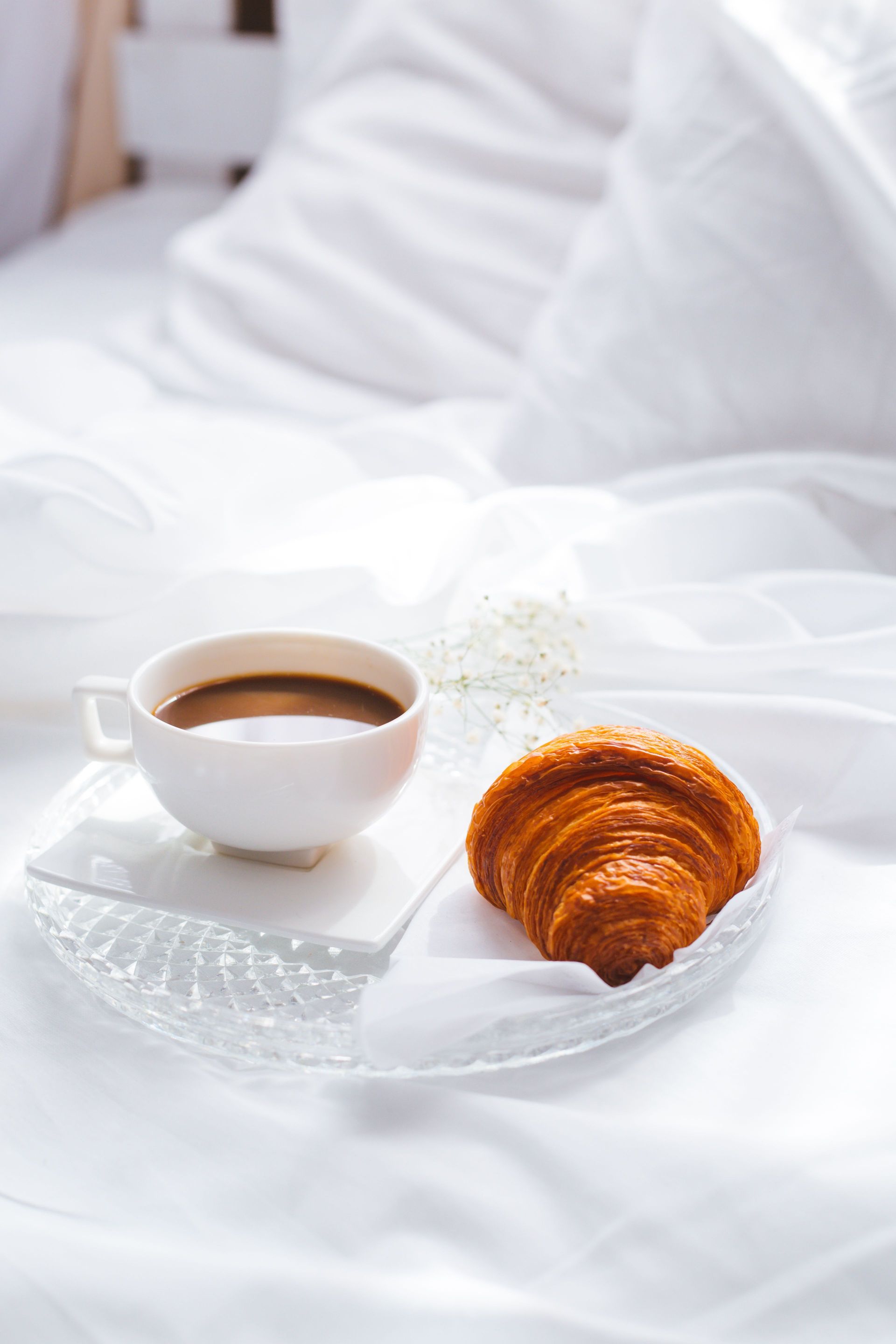 Una tazza di caffè bianco e un croissant su un vassoio trasparente, adagiati su lenzuola bianche stropicciate.