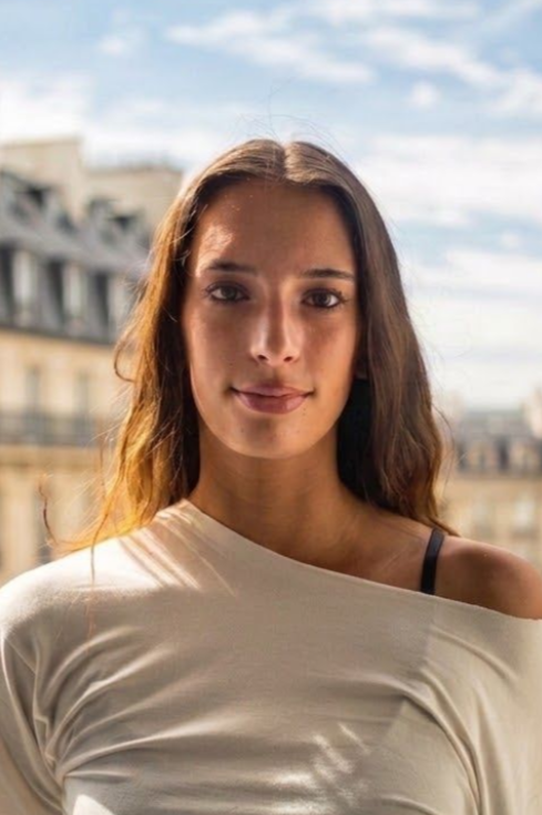 Portrait d'une femme vêtue d'un haut blanc à épaules dénudées, debout à l'extérieur, avec un arrière-plan urbain flou.