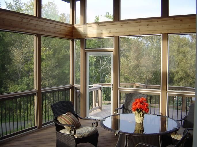 Sunlit screened porch with glass table, black chair, and a vase of orange flowers overlooking trees