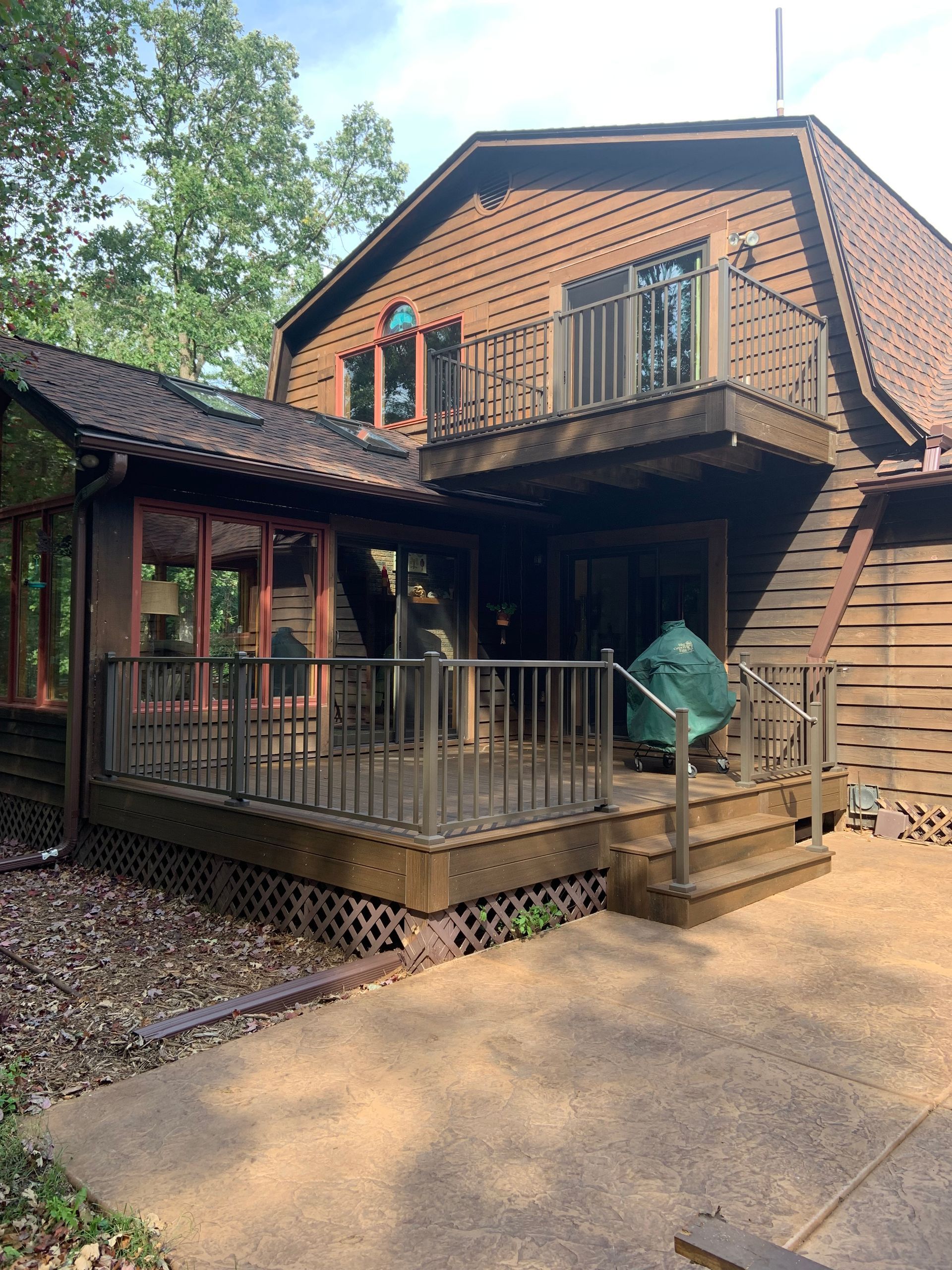 Two-story brick house with wooden deck, stairs, and a green grill on a sunny day