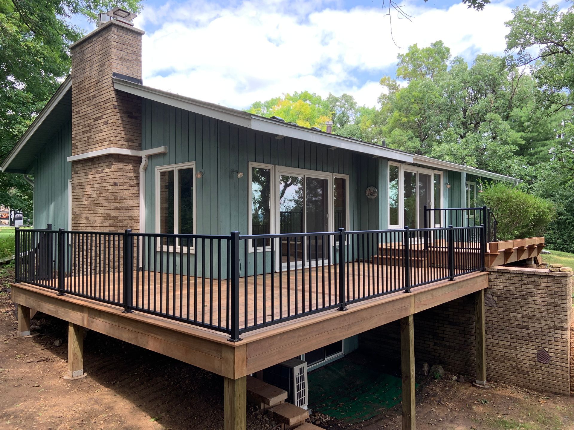 Covered wooden deck beside a dark green house with stone chimney and black railings, surrounded by trees