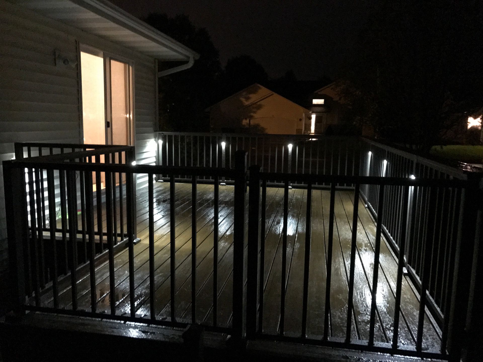 Nighttime backyard deck with black metal railing and wet wooden floor beside a lit house door