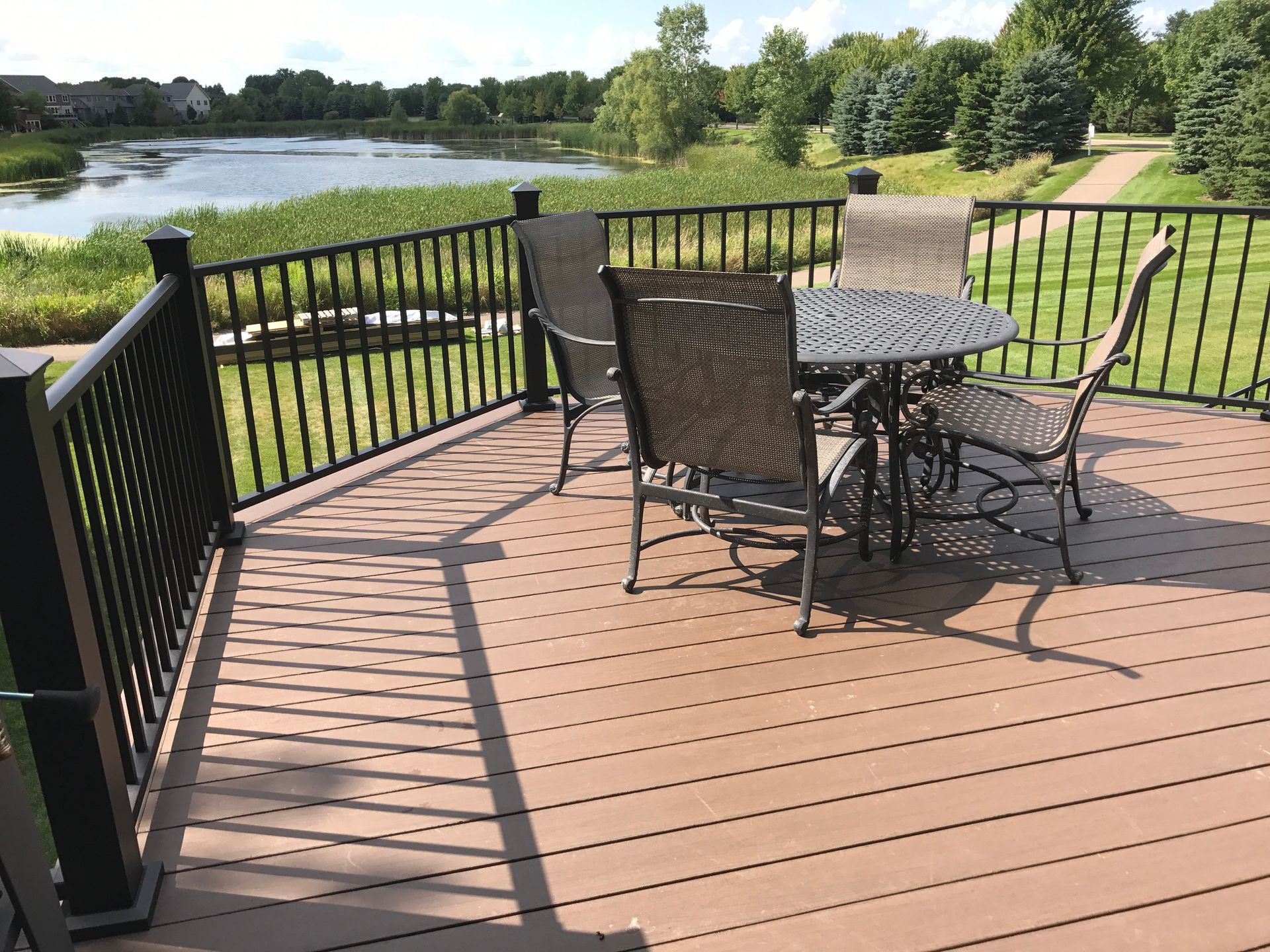Wooden deck with black metal patio table and chairs overlooking a lake and grassy park