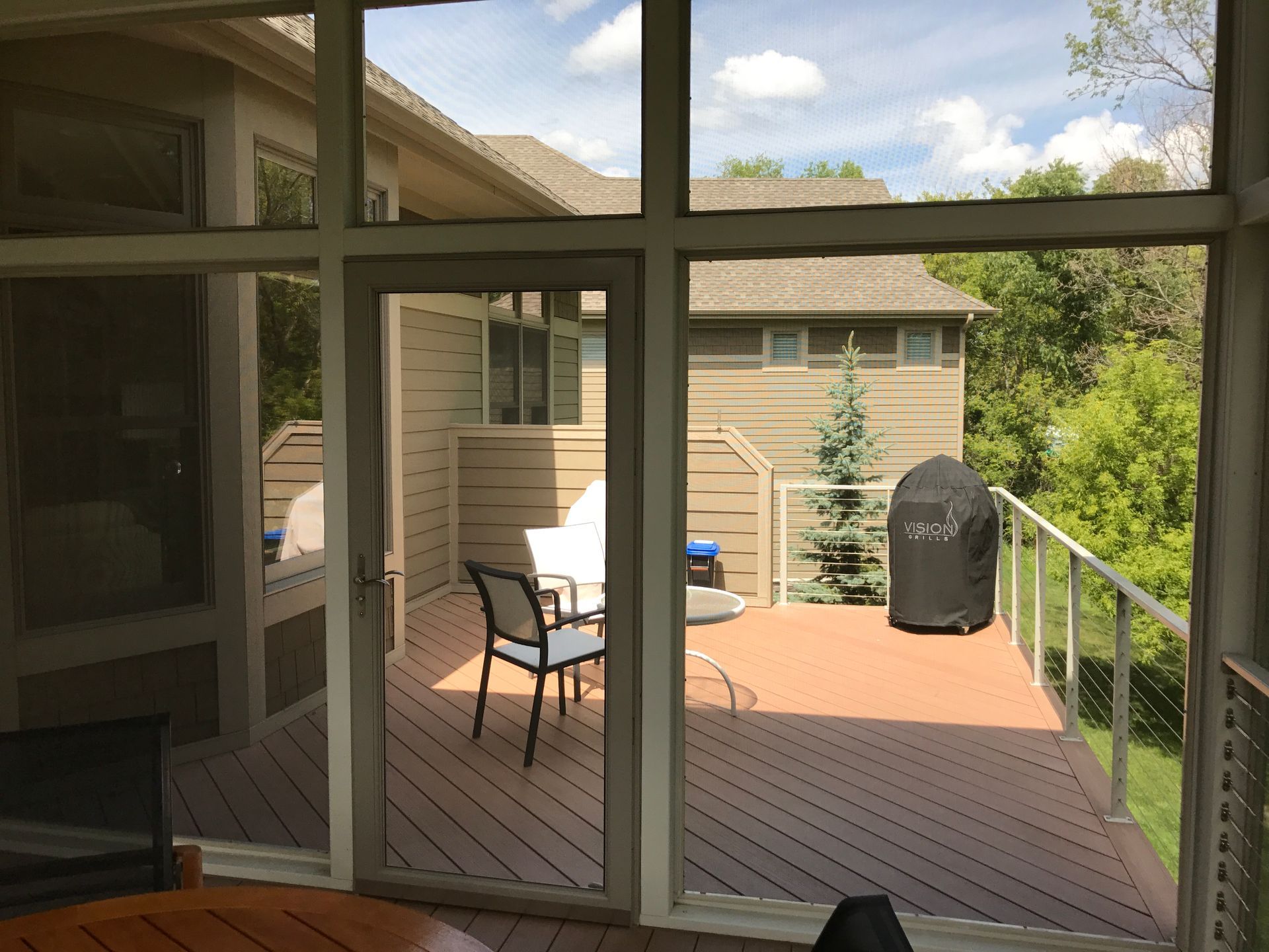 Patio deck with two chairs, grill, and railing viewed through a screened window