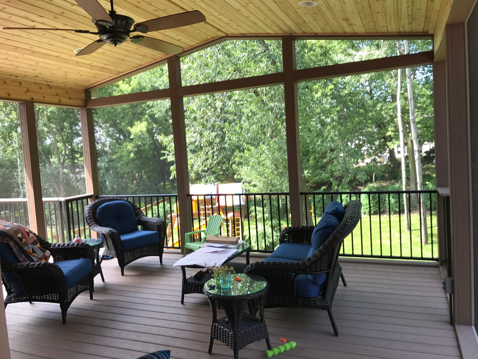 Screened porch with black wicker chairs, small table, and ceiling fan overlooking a green yard