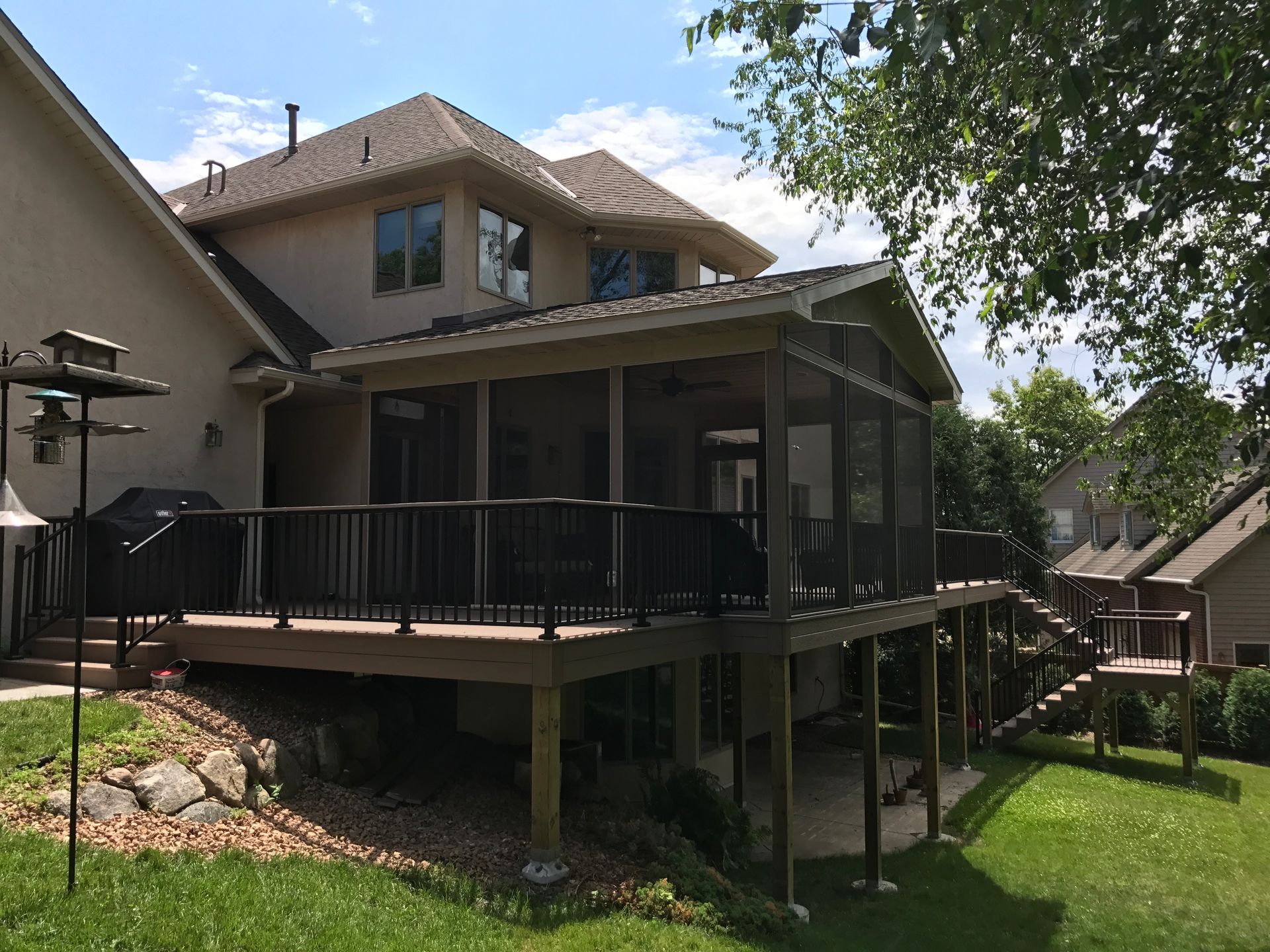 Two-story house with a raised deck, black railing, and sloped backyard under a blue sky