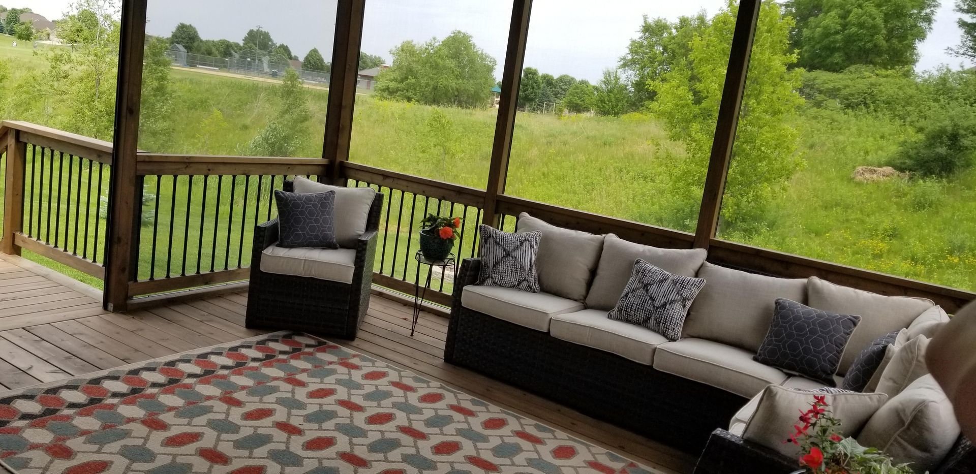 Covered porch with wicker seating, patterned rug, and view of green field beyond the railing.