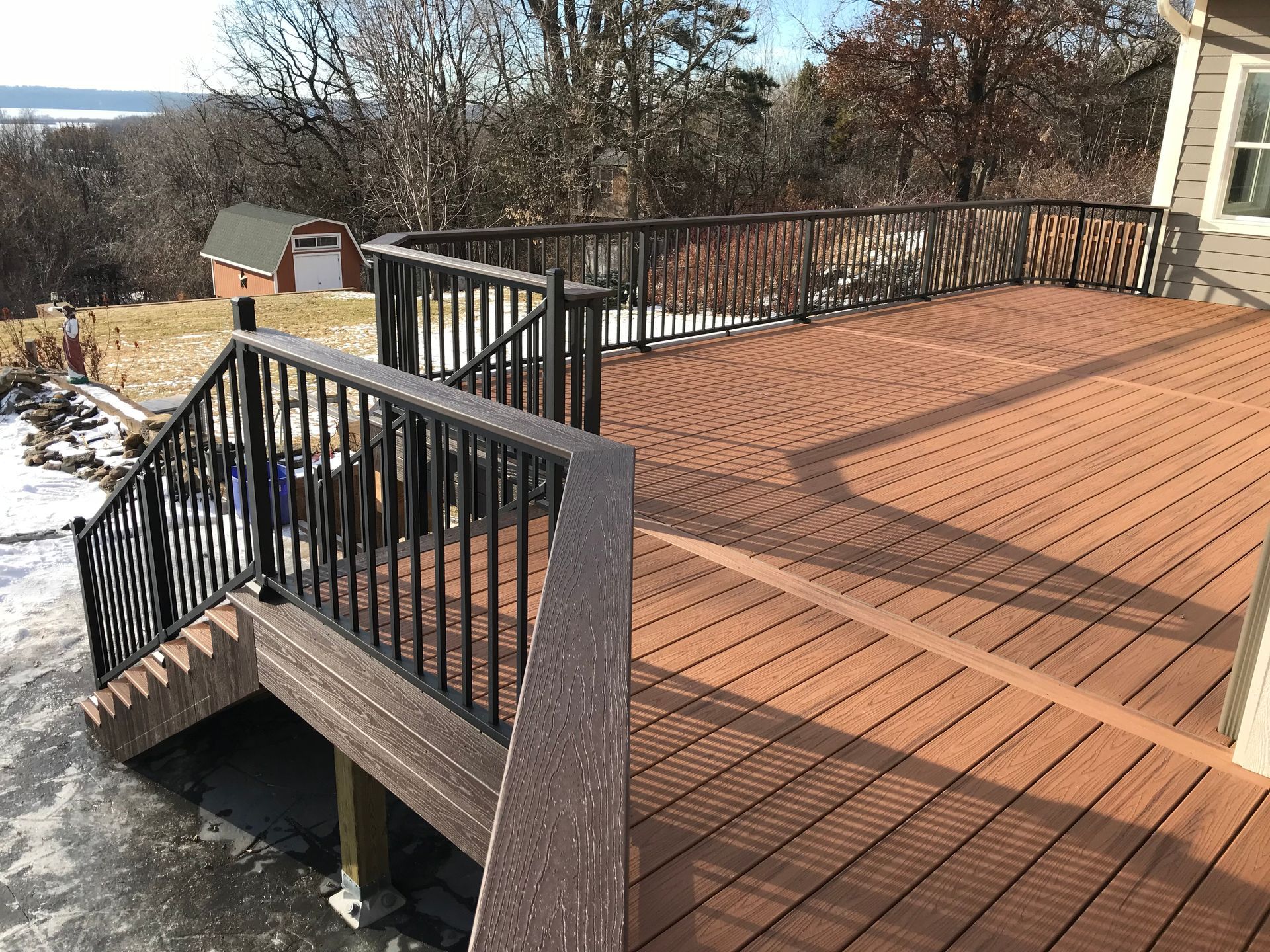 Snowy deck with black railings and stairs overlooking a wooded hillside and small shed
