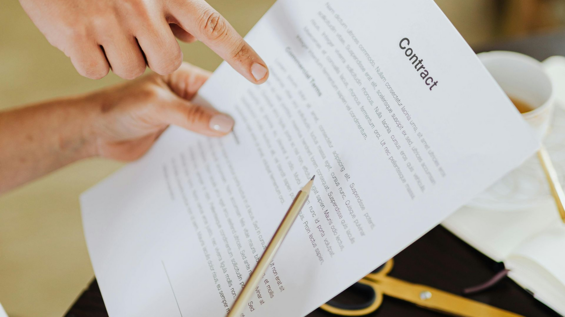 Close-up of hands reviewing a contract document with a pencil, with a coffee cup and gold scissors on a table.