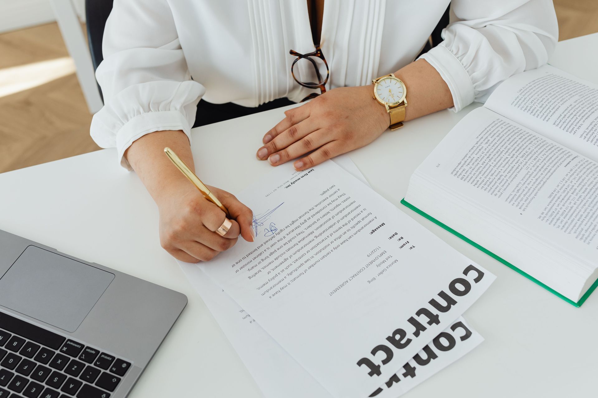 Person writing on paperwork at a desk with a laptop and open documents nearby