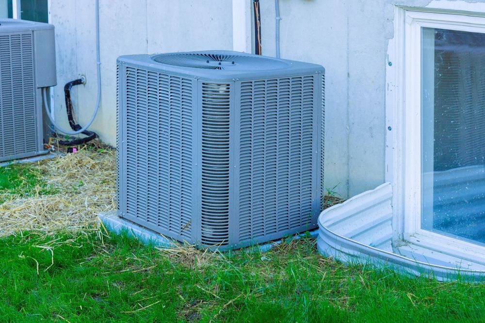 Gray outdoor HVAC unit beside a house wall, with grass and a window well nearby