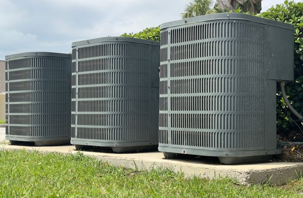 Three gray outdoor HVAC units on a concrete pad beside green grass and bushes