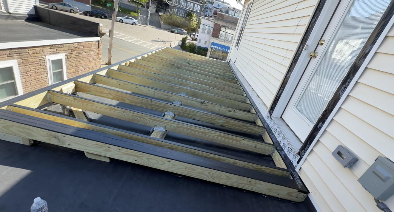 Damaged wooden deck framing beside a white house, viewed from above, with exposed joists and roofing below