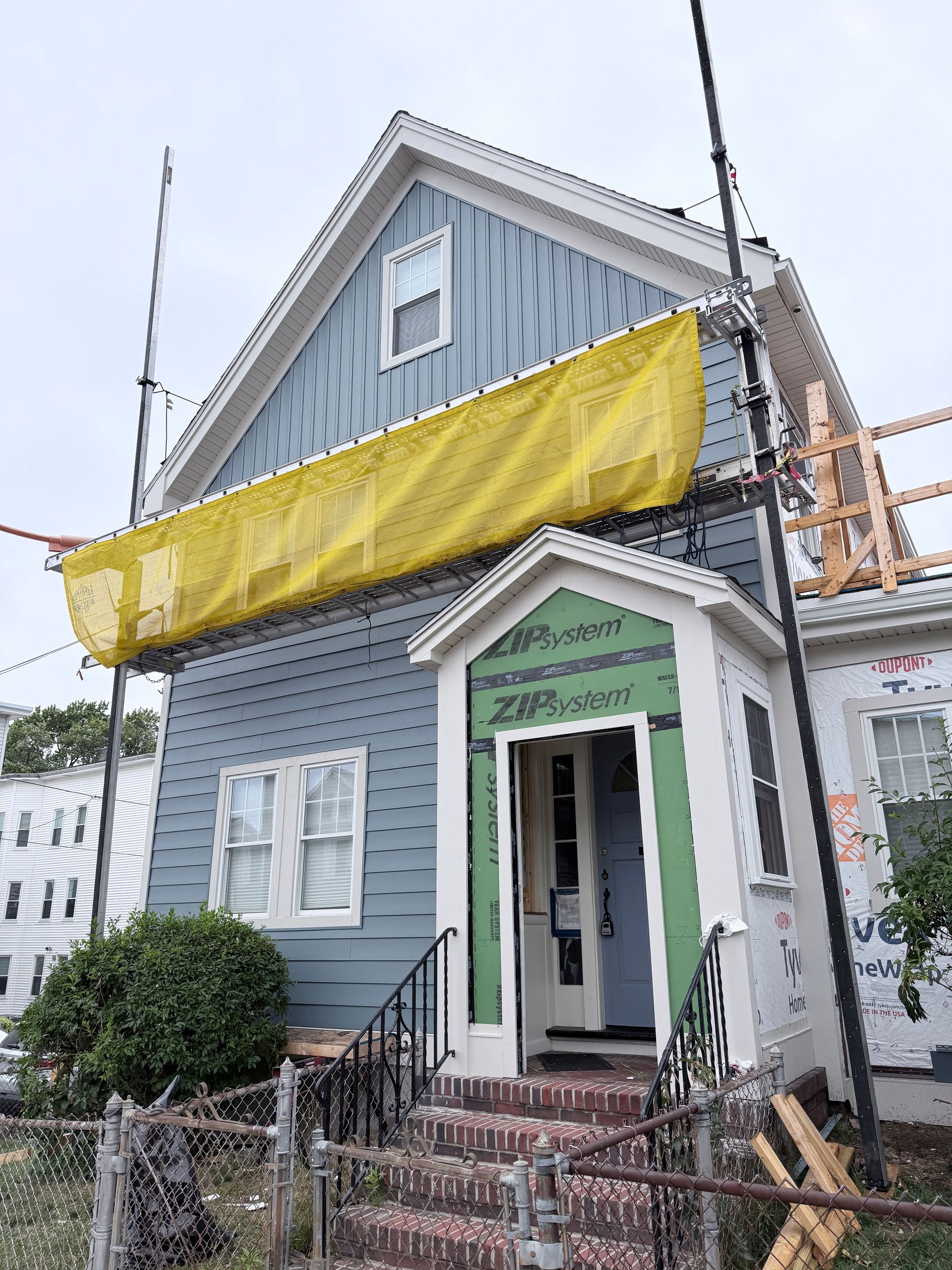 Gray two-story house with white trim, yellow tarp on the upper facade, and a small front porch under renovation