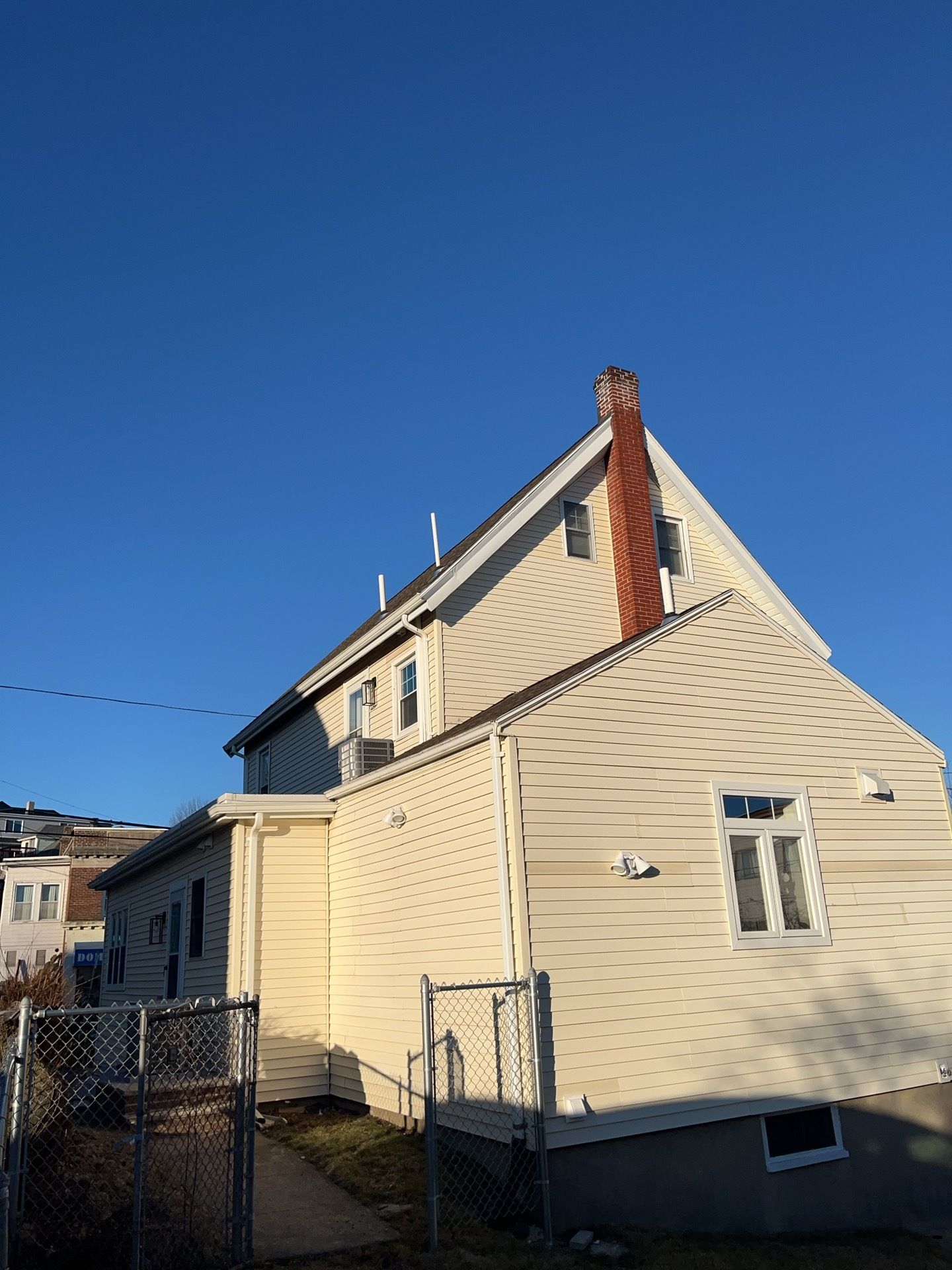 Cream-colored house with a steep roof, brick chimney, and fenced yard under a clear blue sky