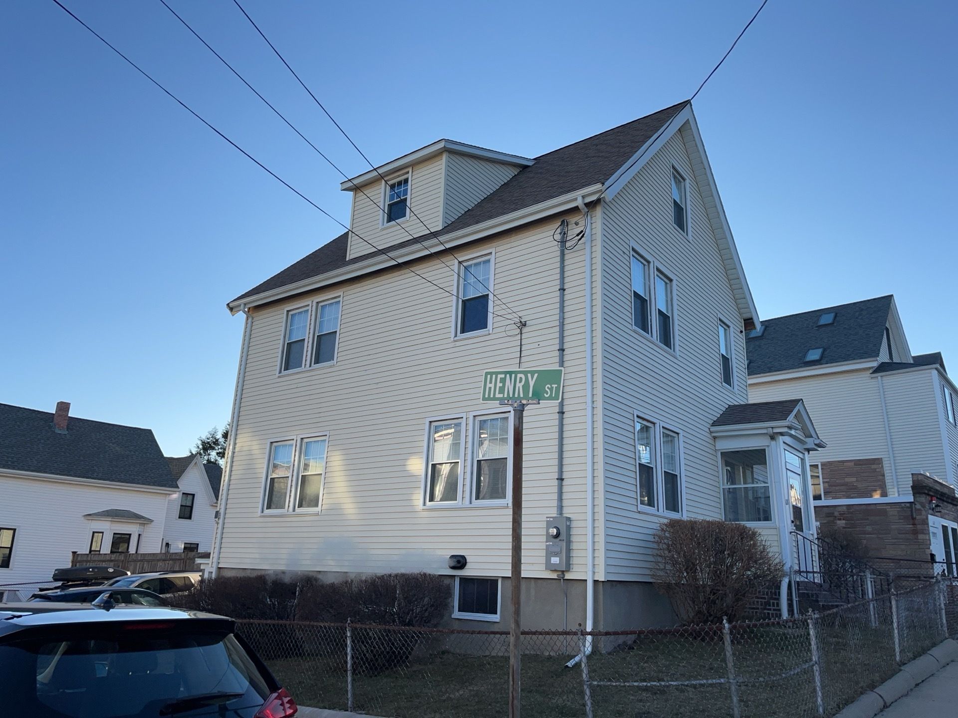 Two-story beige house on a street corner under a clear blue sky, with parked cars and a chain-link fence.