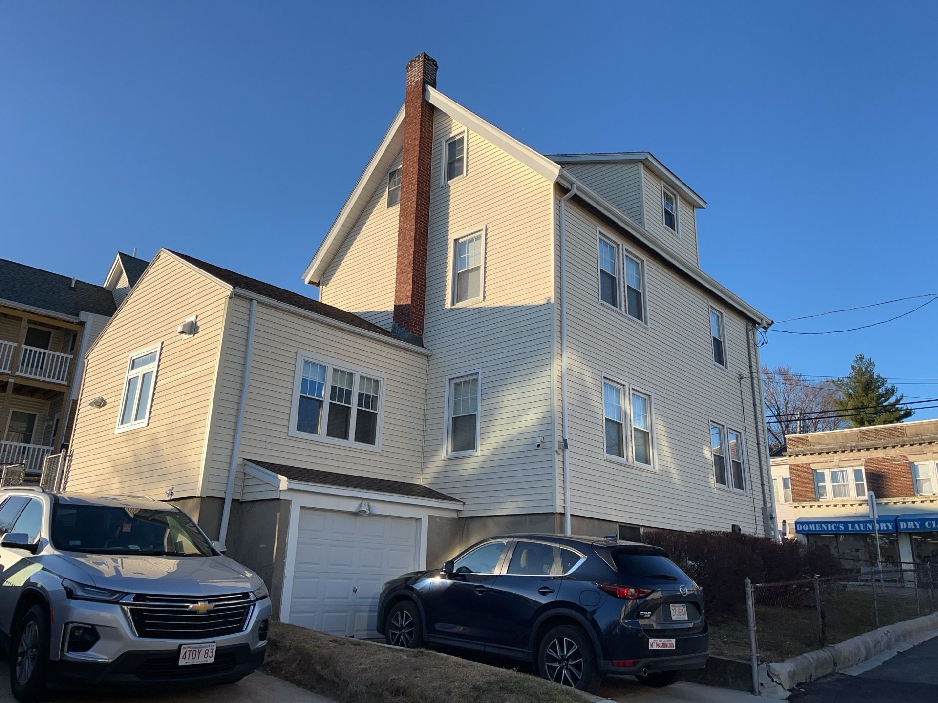Two-story beige house with parked cars in the driveway on a sunny day