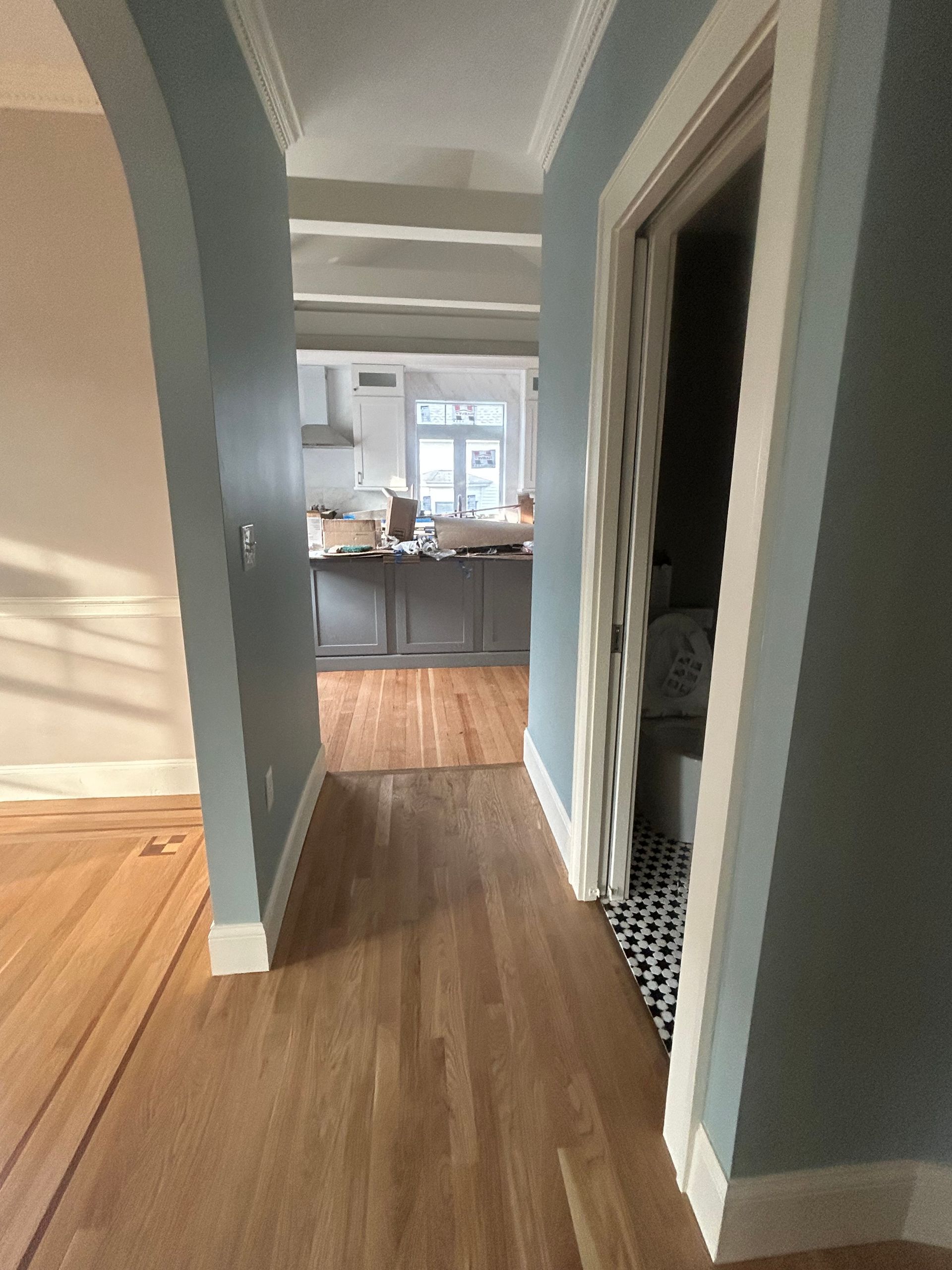 Narrow hallway with wood floors and gray walls leading to a bright kitchen.