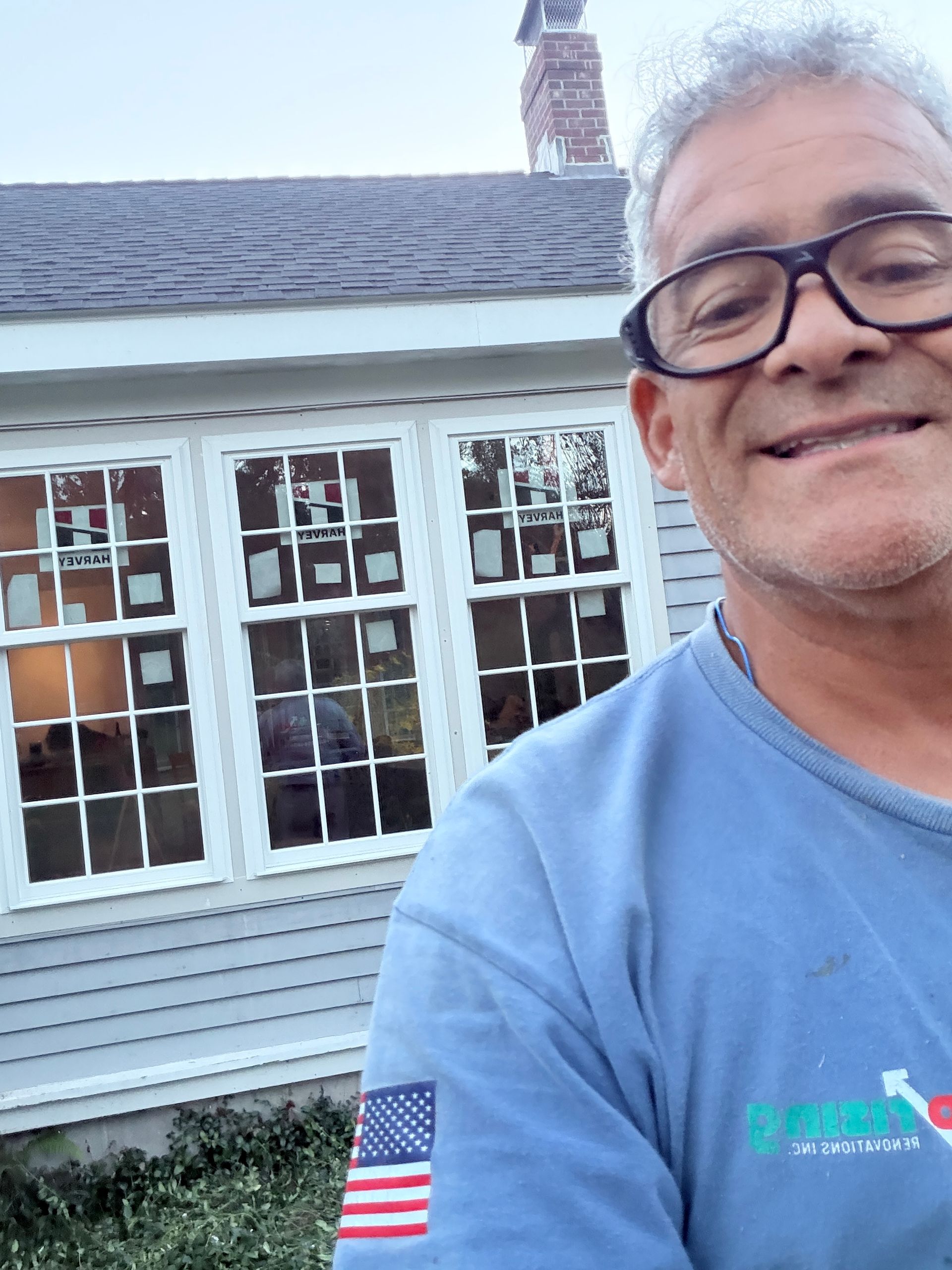 Man smiling in a light blue shirt taking a selfie in front of a house with white-trimmed windows.