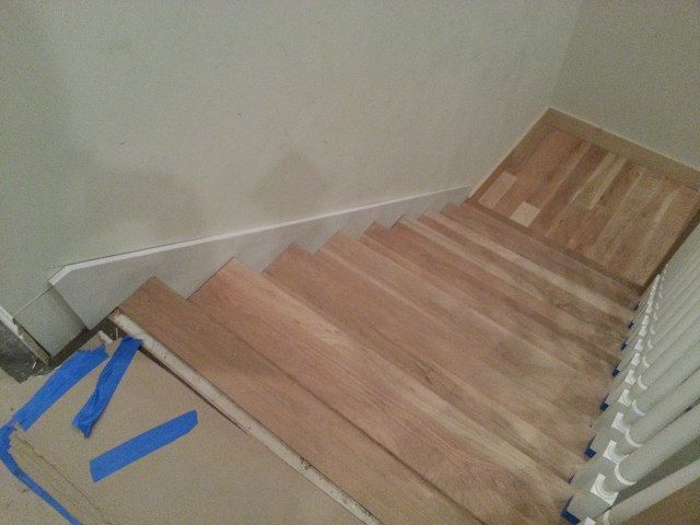 Staircase with wood flooring and white railing, viewed from above, with blue tape marking the bottom step area