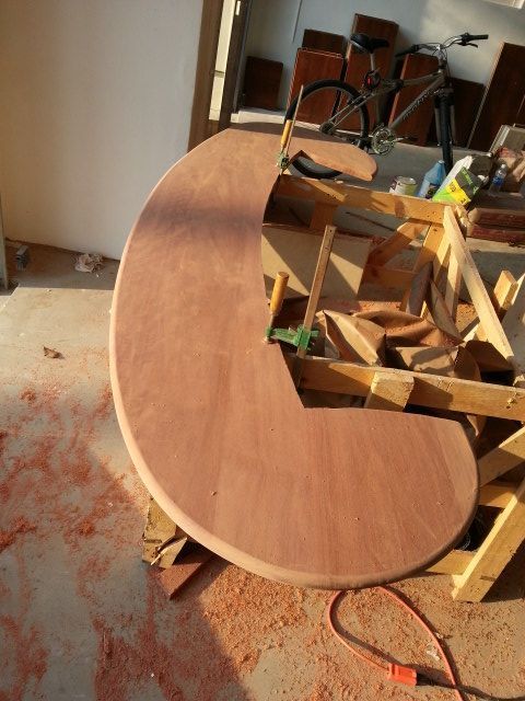 Curved wooden tabletop under construction in a workshop, with clamps and lumber around it.