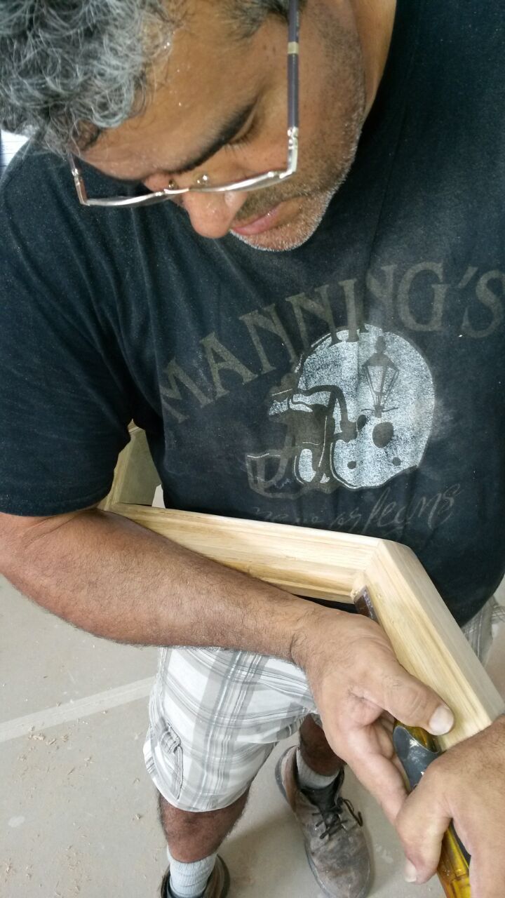 Person holding a wooden frame while using a tool, wearing glasses and a black T-shirt.
