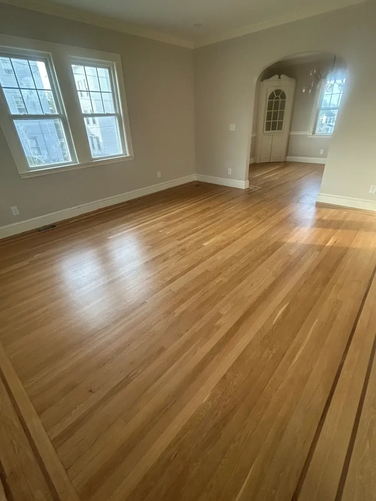 Empty sunlit room with hardwood floors, beige walls, and windows on the left.