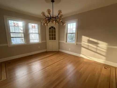Empty sunlit dining room with hardwood floors, beige walls, chandelier, and three windows