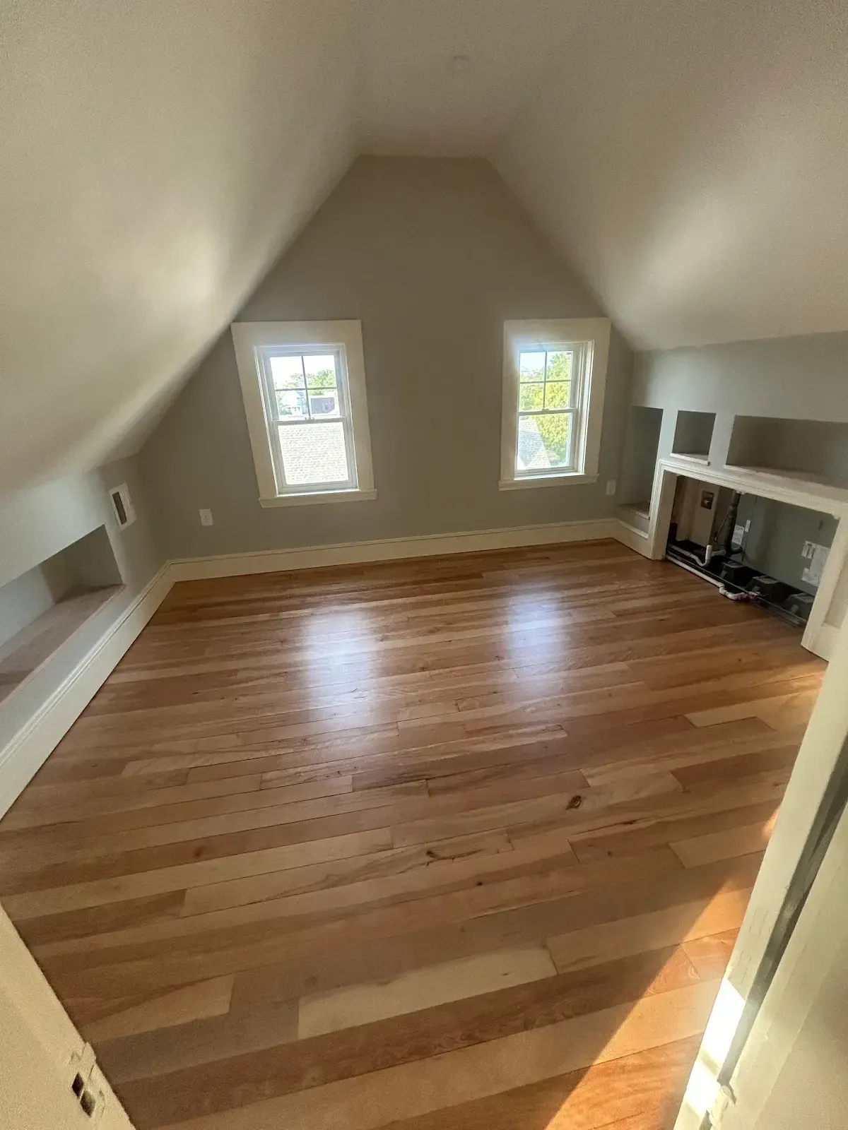 Empty attic room with hardwood floor, two windows, and built-in shelving on the right