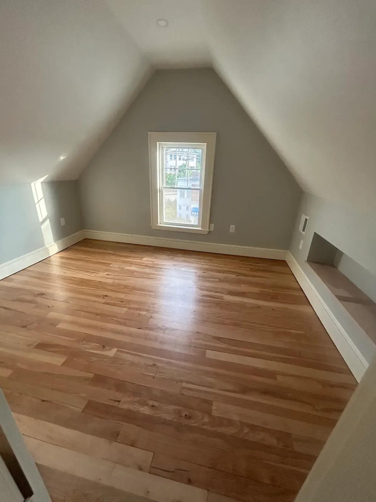 Empty attic room with sloped white walls, hardwood floor, and a small central window.
