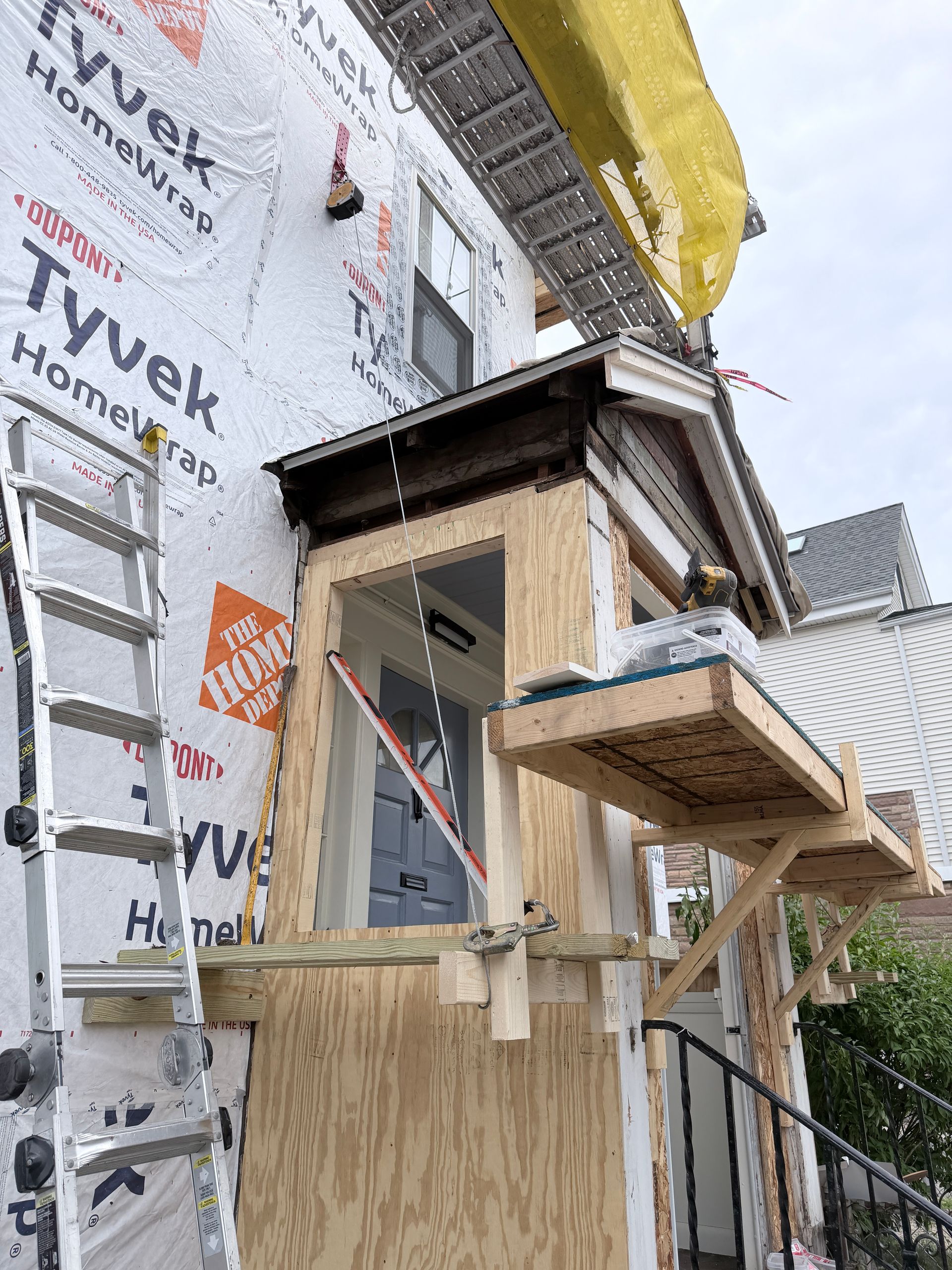 Partially built house with plywood porch, wrapped in Tyvek, and a yellow tarp on the roof