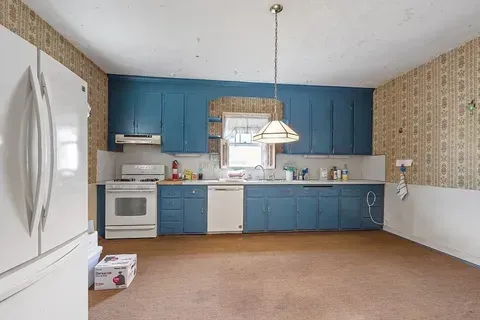 Empty kitchen with blue cabinets, white appliances, and beige wallpaper under a hanging light fixture