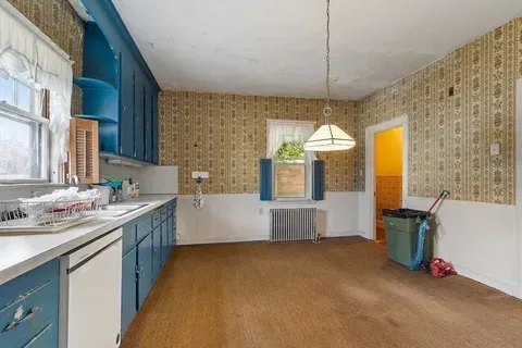 Empty kitchen with blue cabinets, patterned wallpaper, a hanging light, and a trash bin by the door.