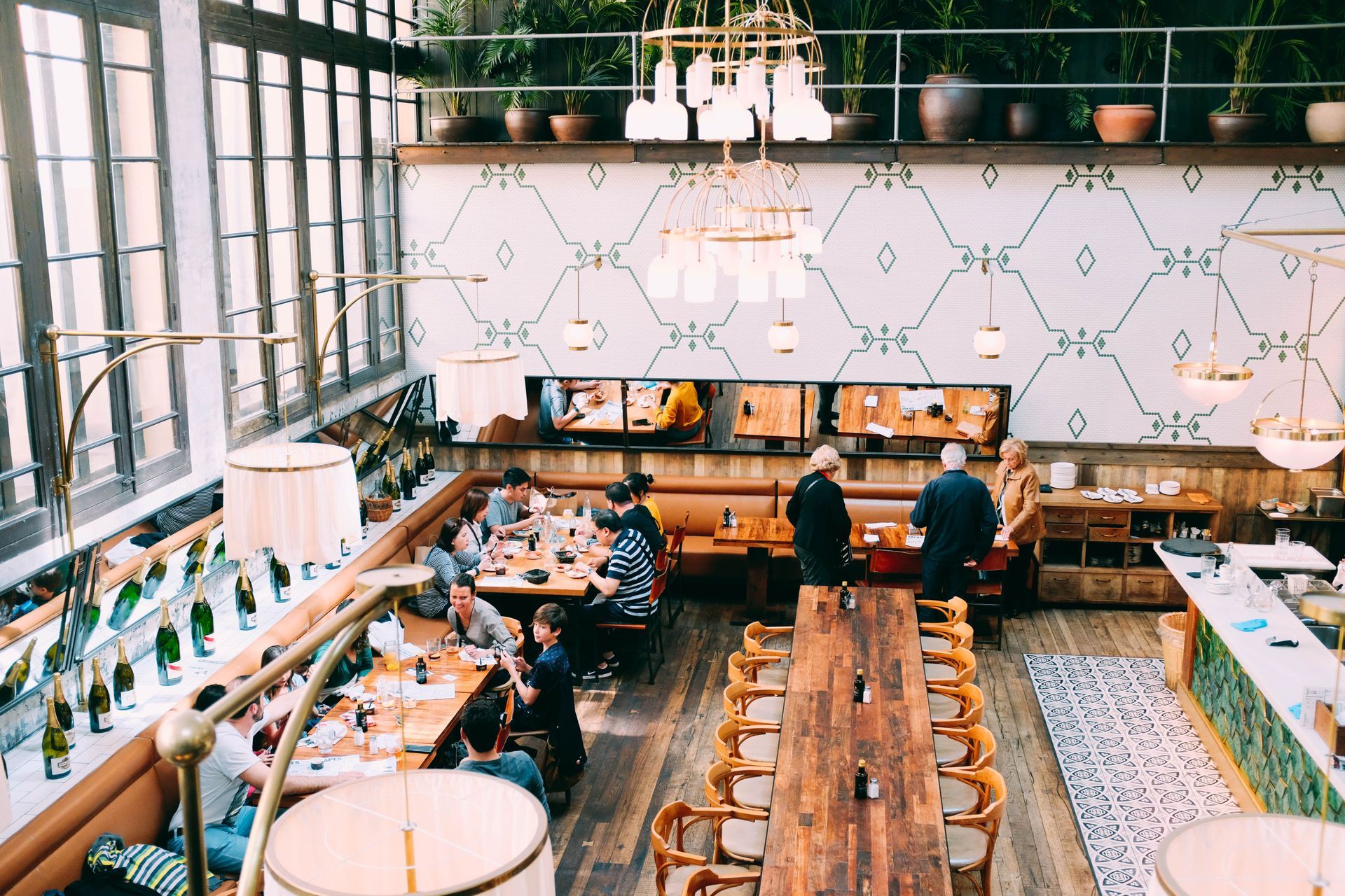 Bright café interior with long wooden tables, hanging lights, and patrons seated near large windows.
