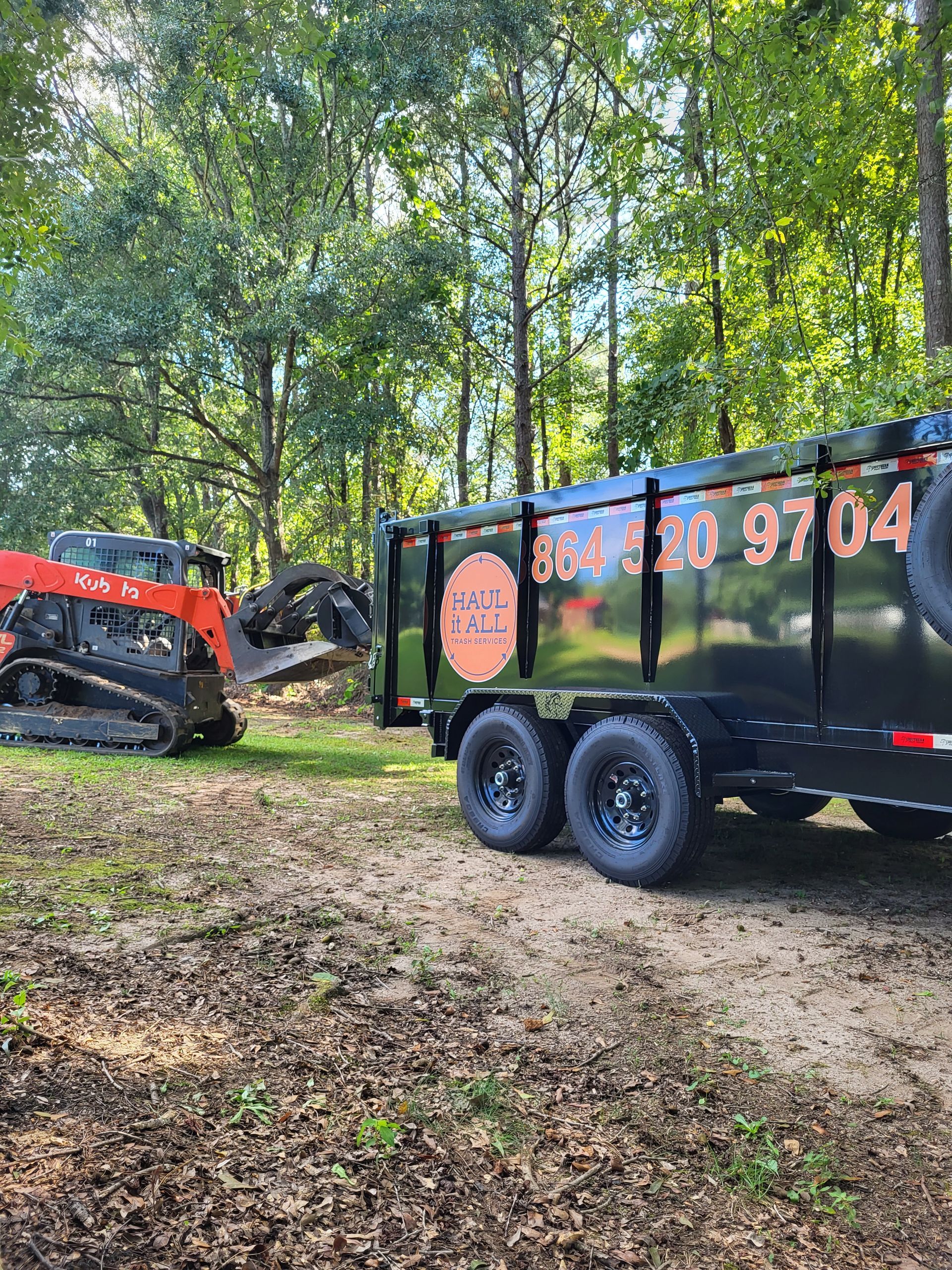 Orange skid steer beside black dump trailer in a wooded area, hauling leaves and debris.