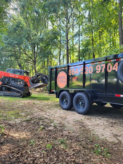 Black landscaping trailer beside orange skid steer in a wooded area, with phone number on the trailer.
