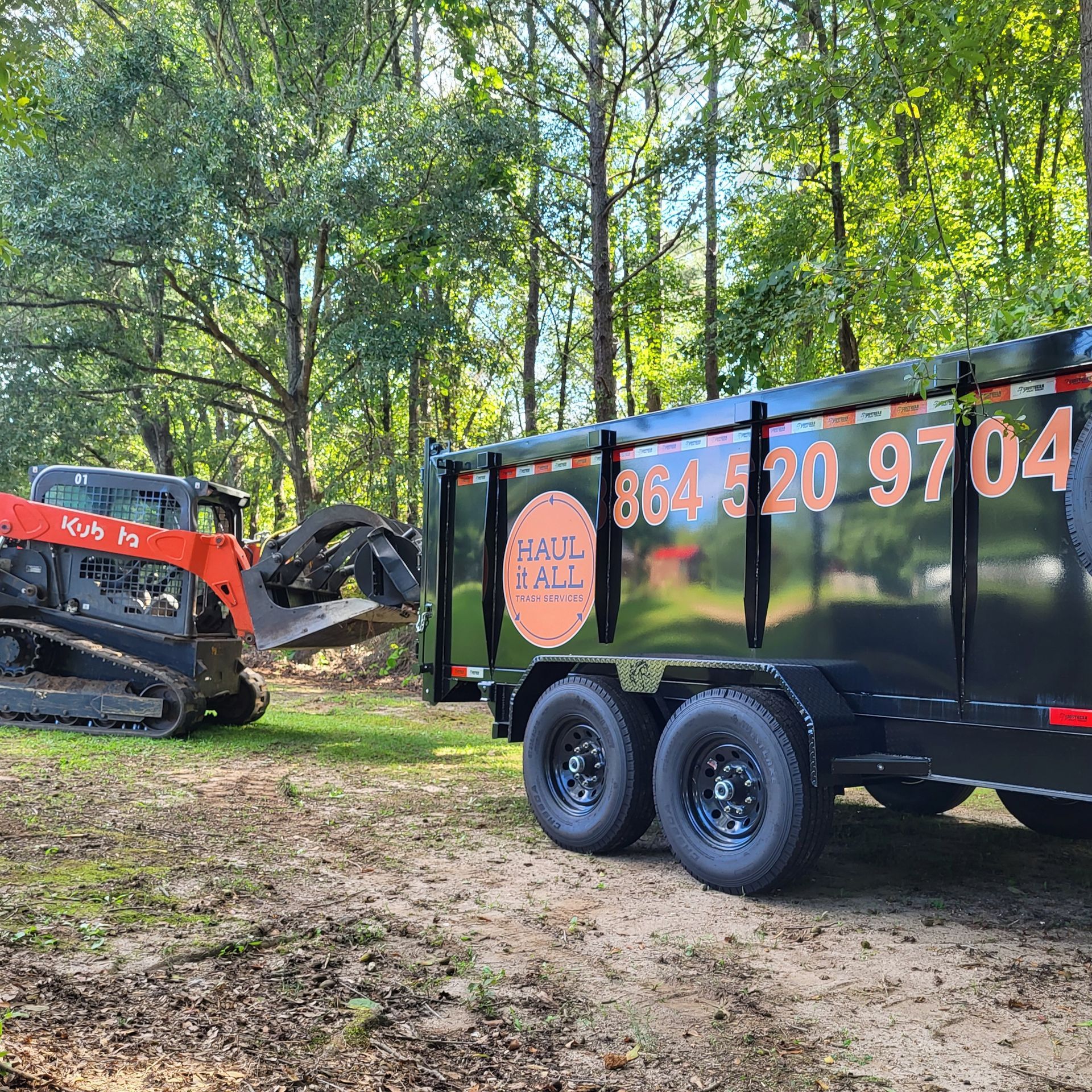 Red skid steer loading debris into a black landscaping trailer in a wooded area