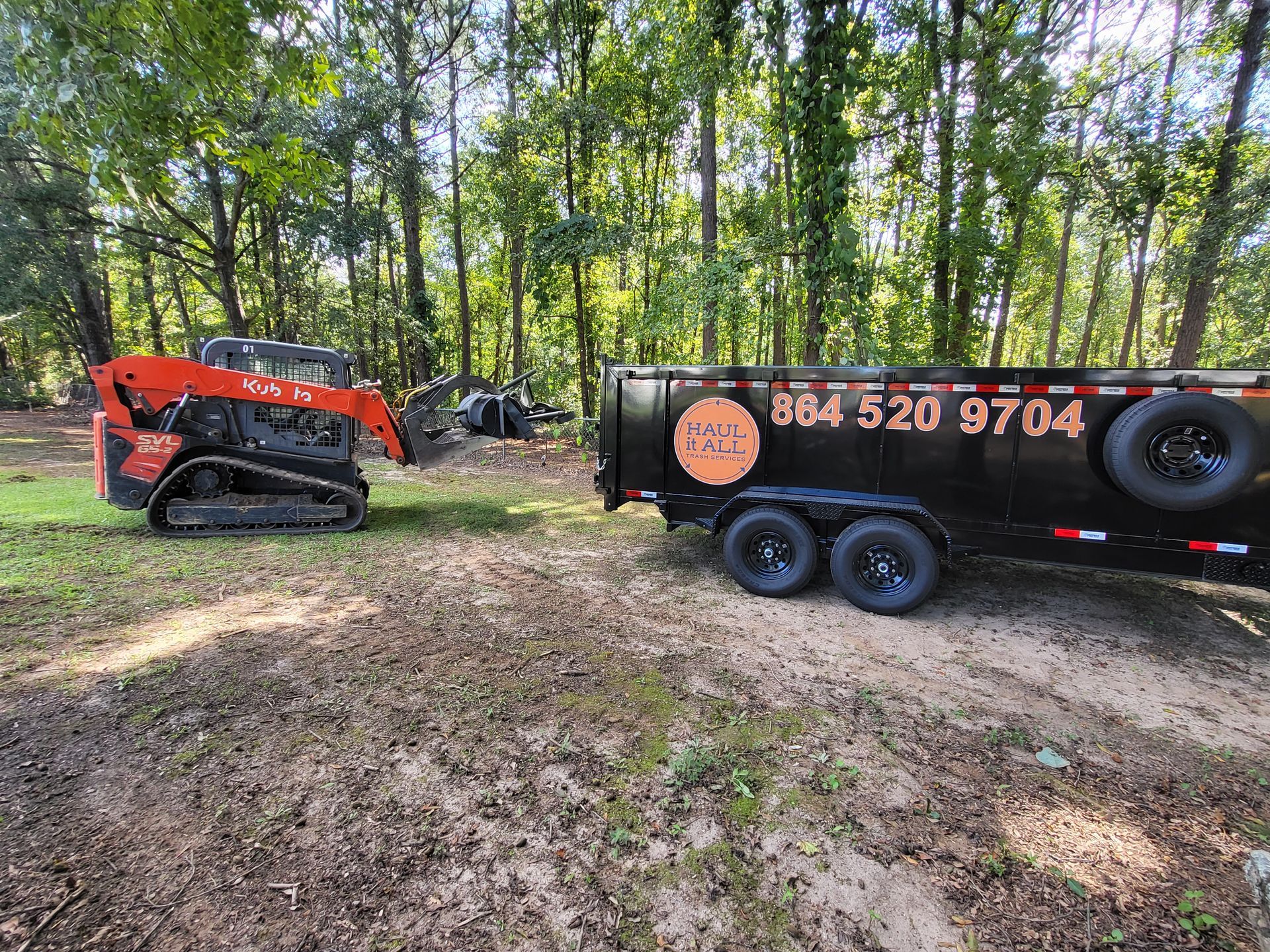 Orange skid steer beside black trailer in a wooded clearing, with another machine in the background.