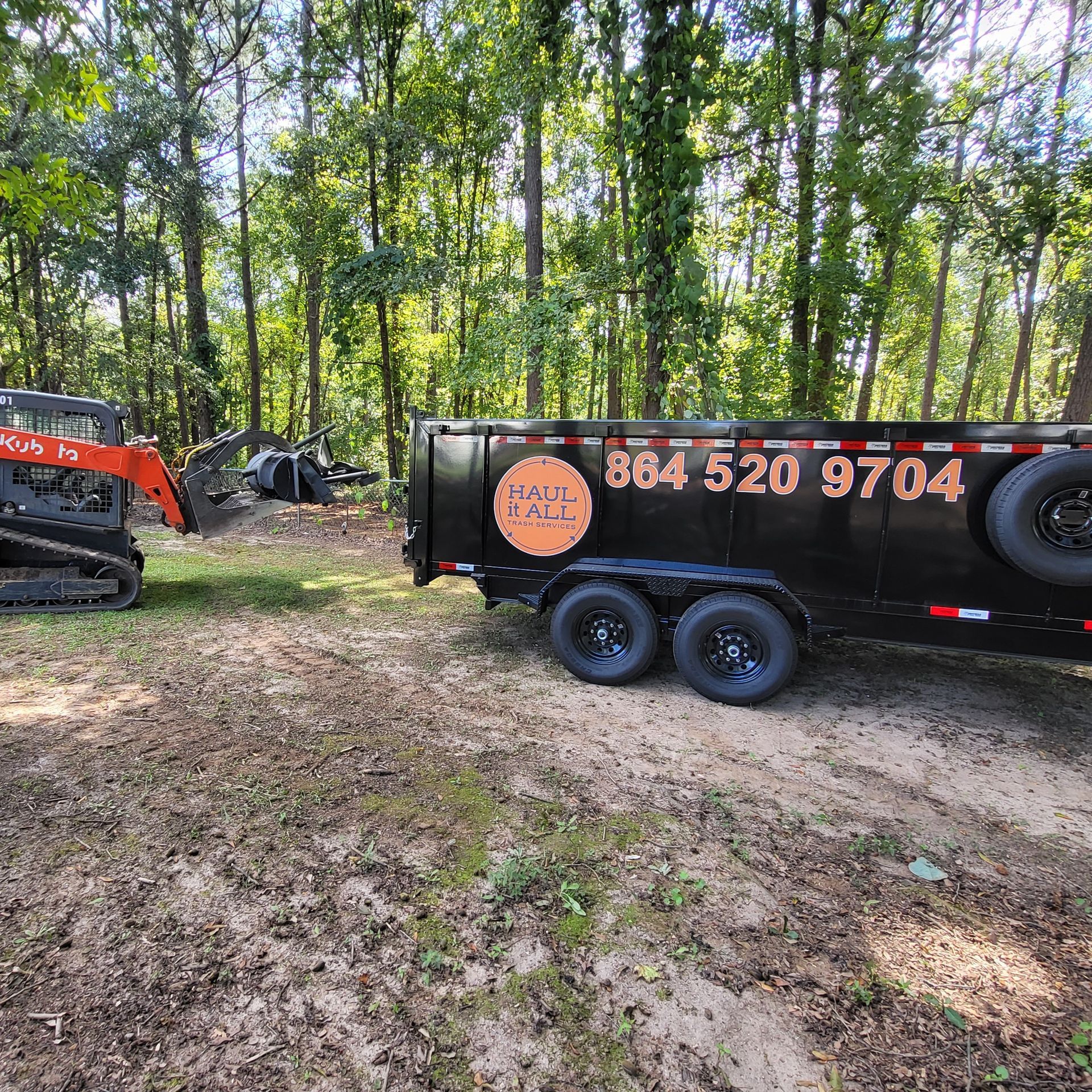 Black utility trailer with phone number in wooded clearing beside an orange excavator