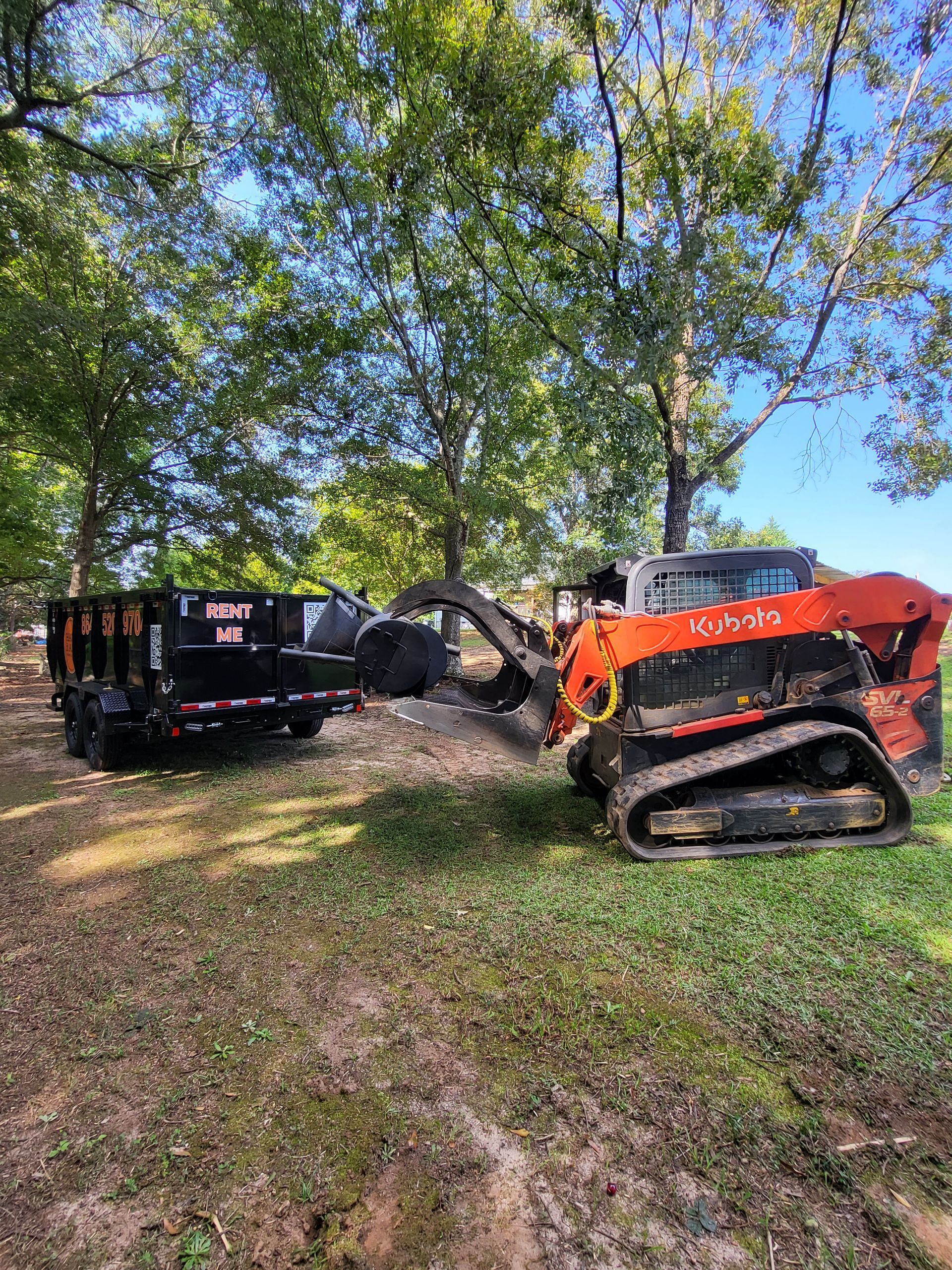 Orange excavator beside a tipped black trailer under trees in a grassy field