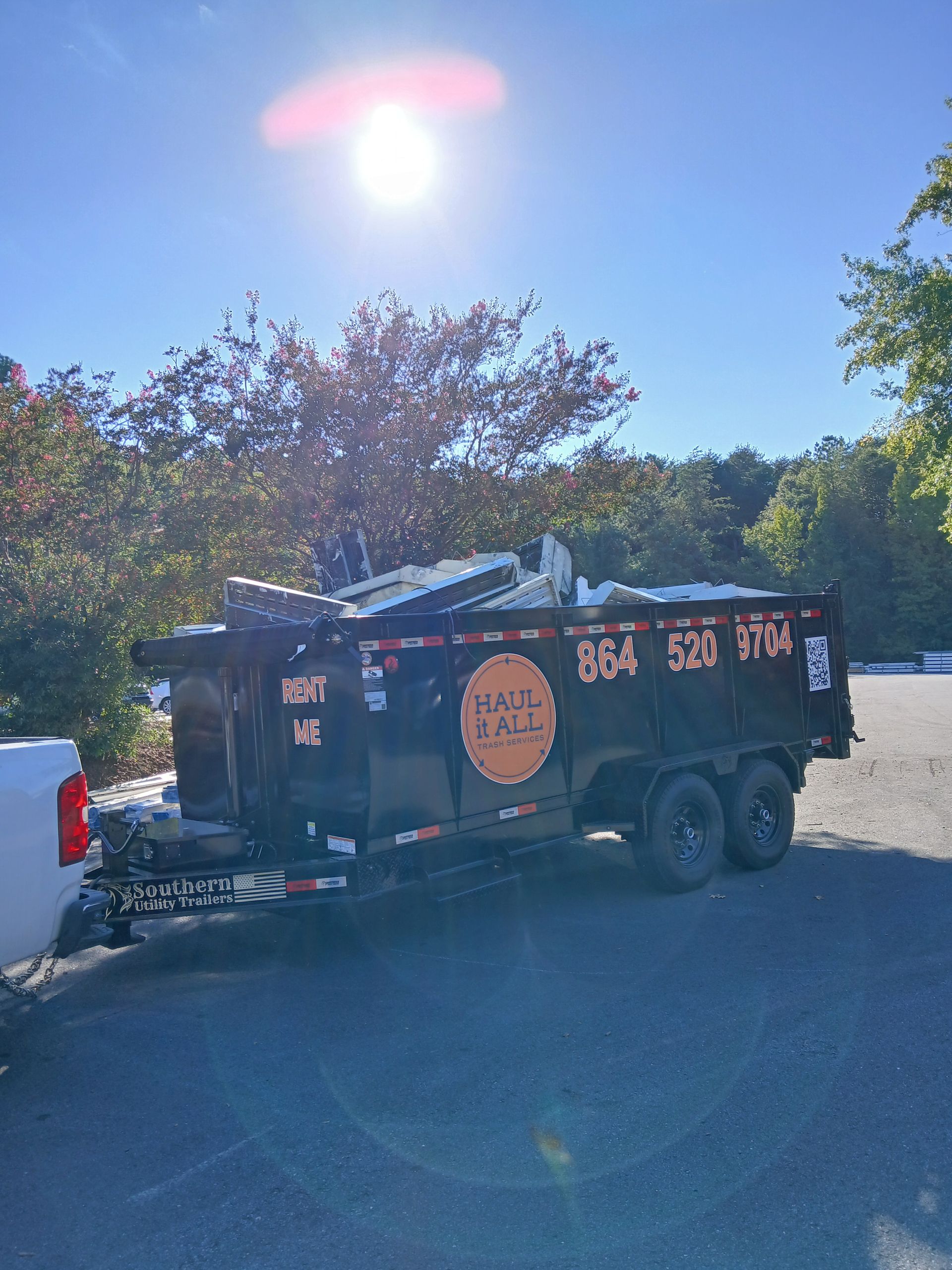 Black utility trailer with signage parked on a sunny roadside beside trees and a white truck