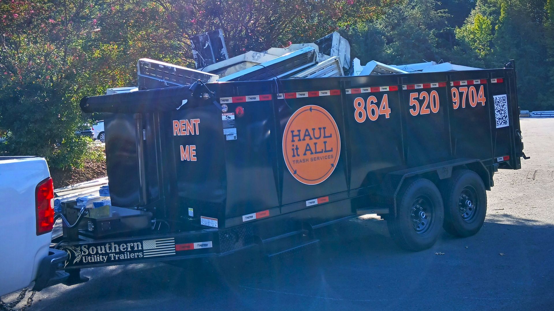 Black Hall & Hall dumpster trailer parked outdoors on a sunny roadside, surrounded by trees.