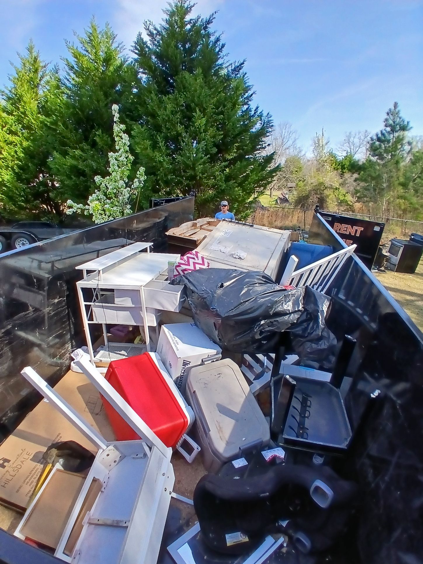Pile of mixed trash and broken furniture piled outdoors beside green trees.