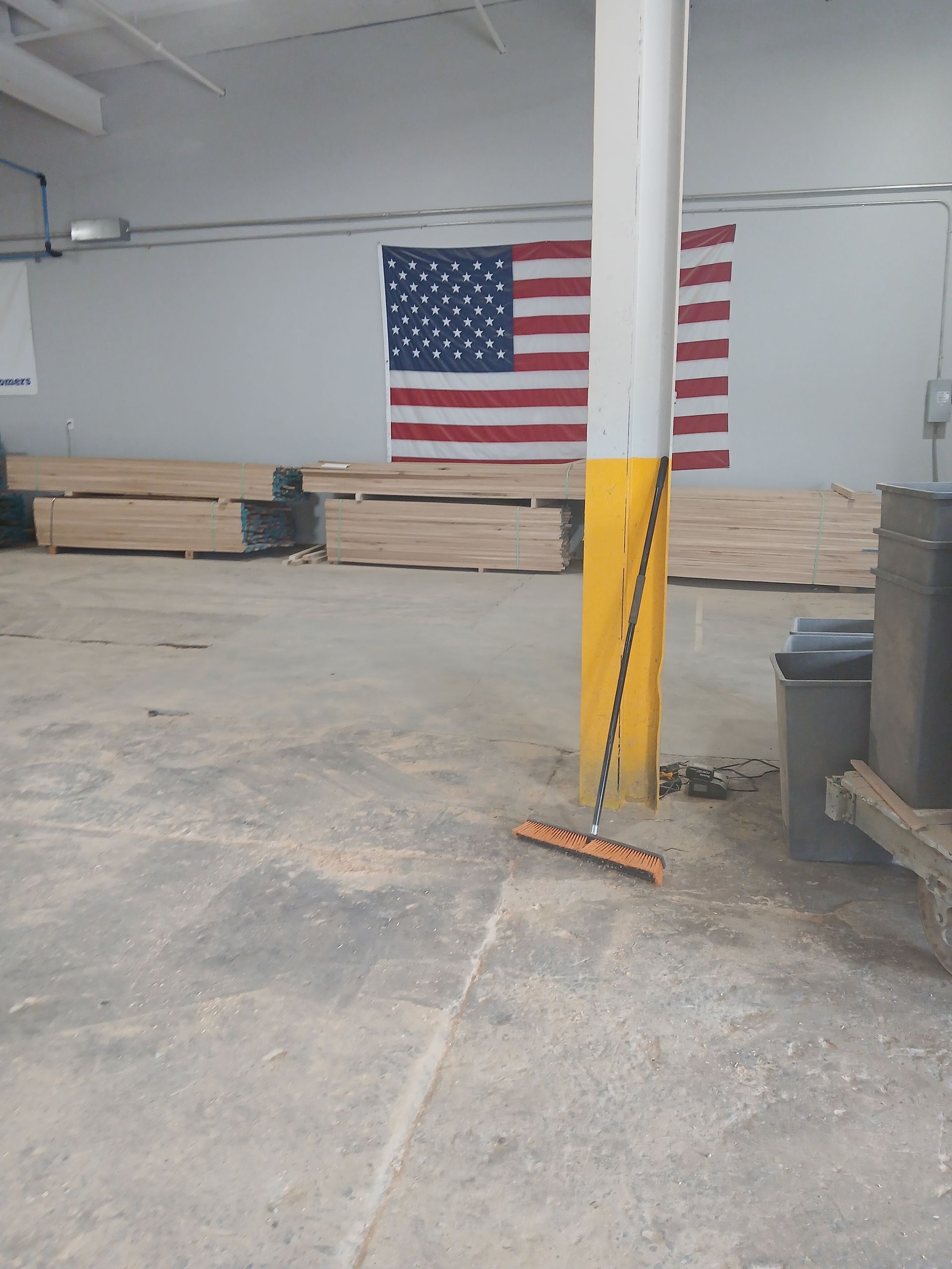 Warehouse interior with stacked wood planks, an American flag, and a broom leaning against a yellow column.