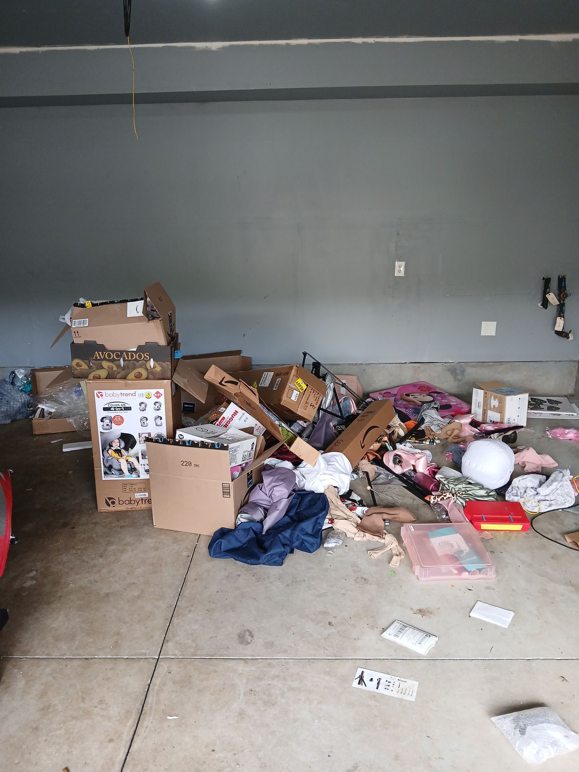 Messy garage floor covered with scattered boxes, trash, and clothing against a gray wall.