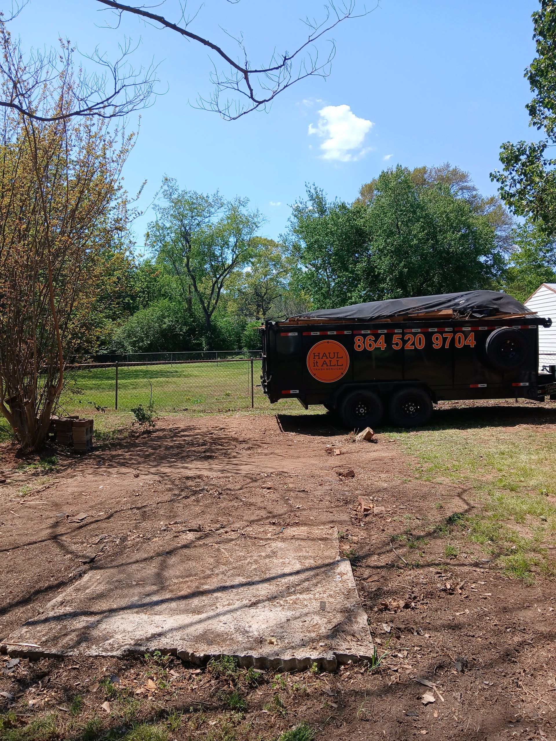 Small black trailer with orange lettering parked on a dirt path in a sunny grassy yard, surrounded by trees.