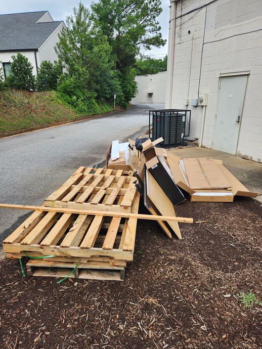 Wooden pallets and cardboard stacked beside a white building on a gravel driveway