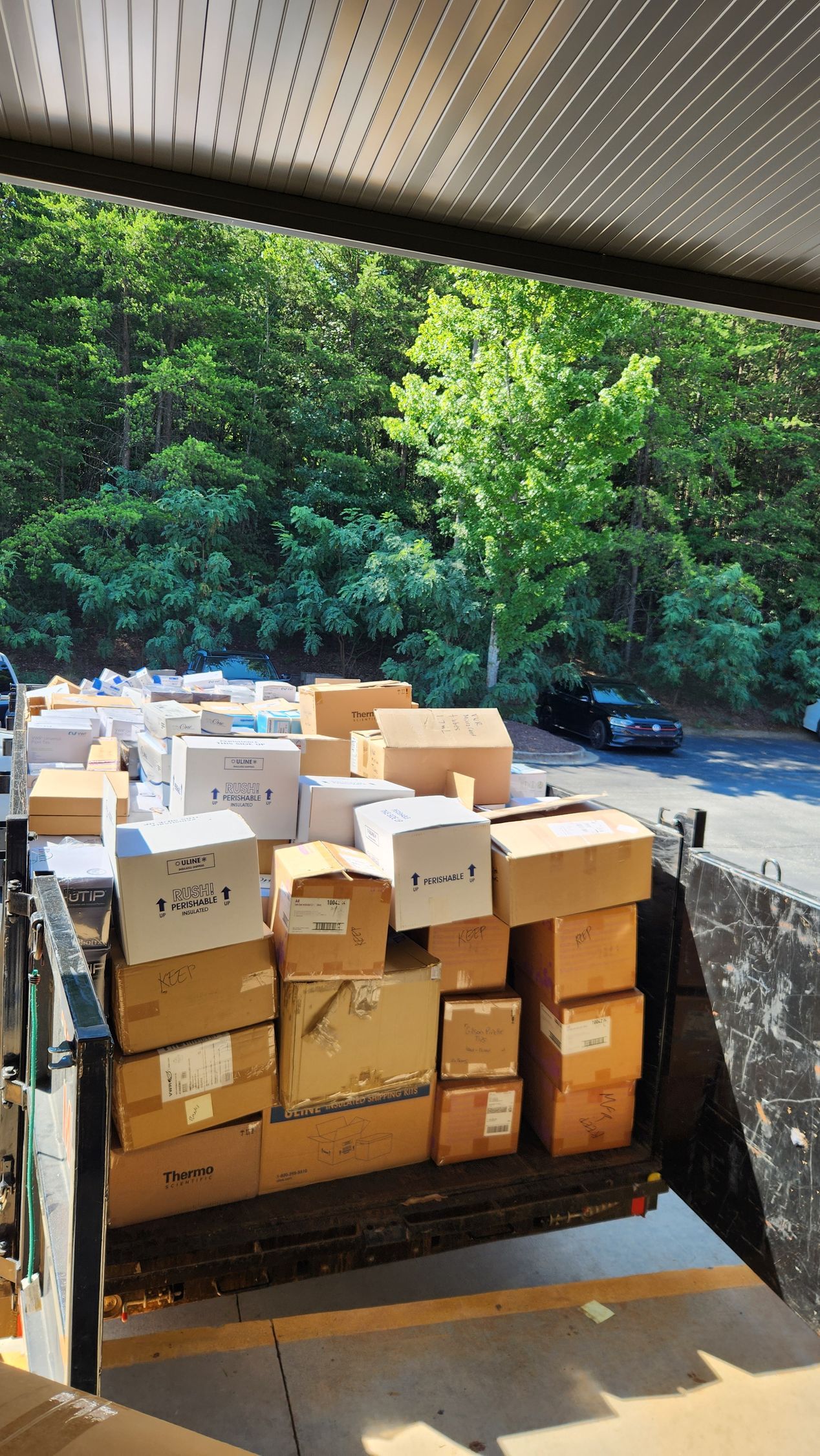 Boxes of assorted packages stacked on a flatbed truck under a covered loading area, with trees in the background